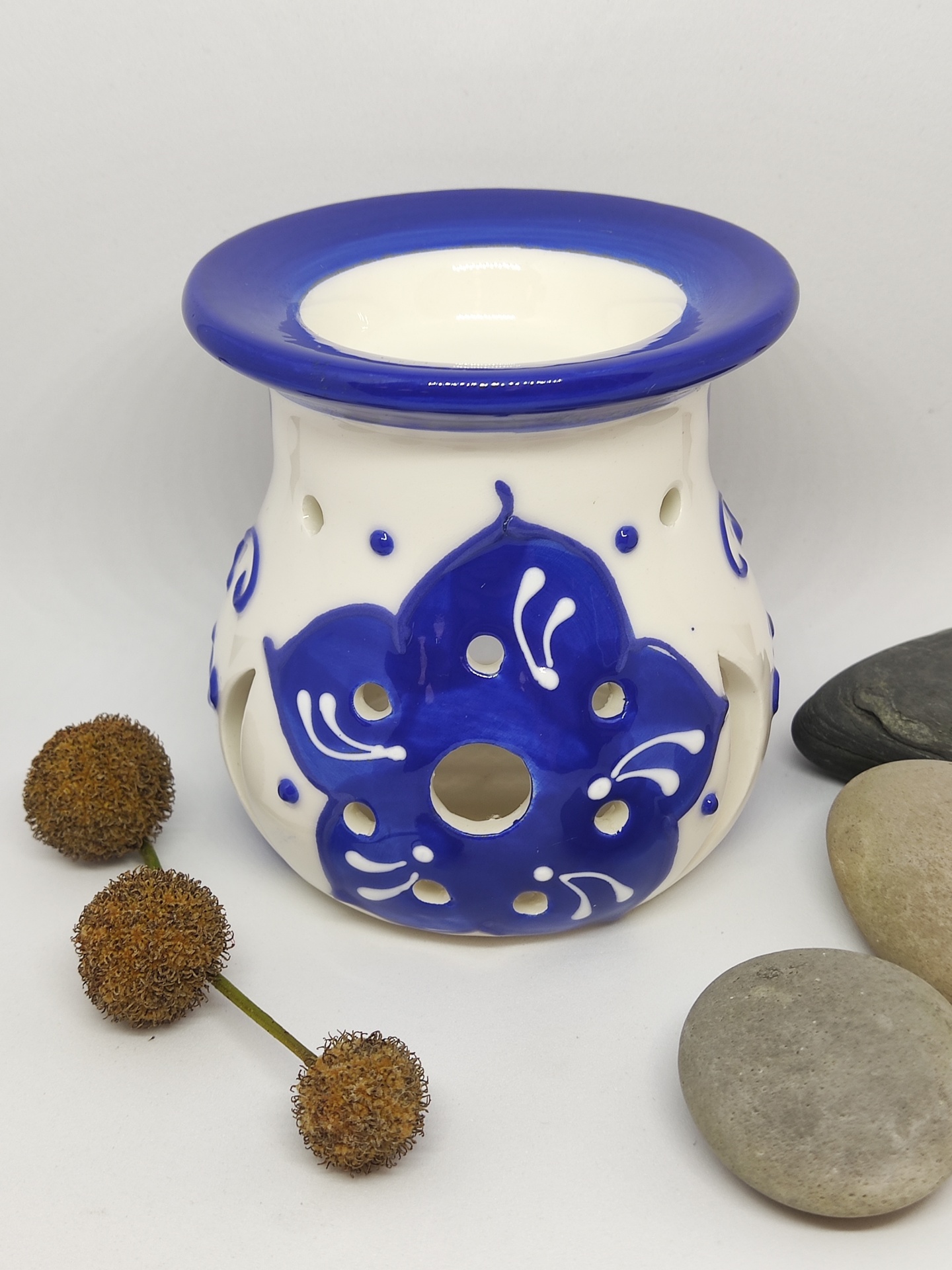 Queimador de cerâmica branco com decoração azul em forma de flor, rodeado por pedras e bolbos secos
