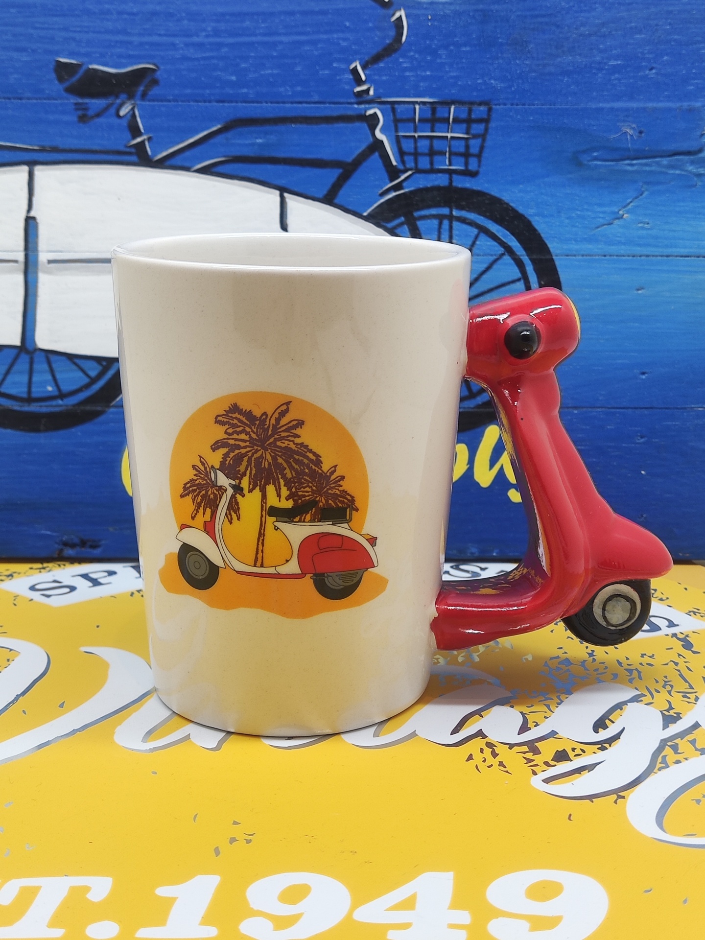 Caneca branca com pega em forma de scooter vermelha e desenho de scooter com palmeiras.