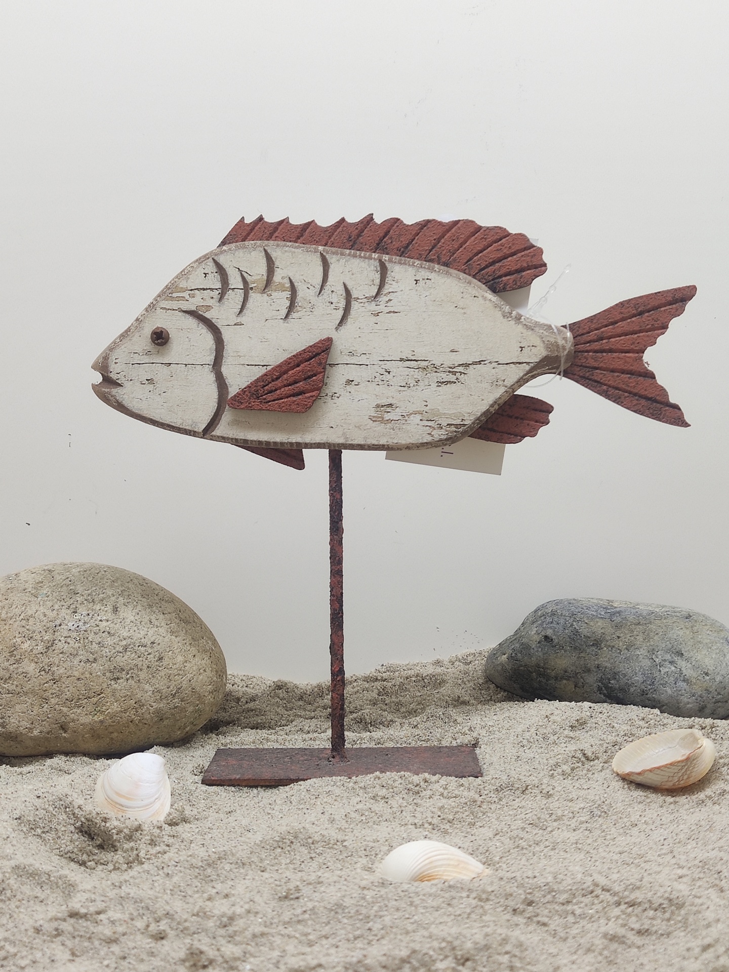 Figura decorativa de peixe em madeira com suporte metálico em areia com pedras e conchas.