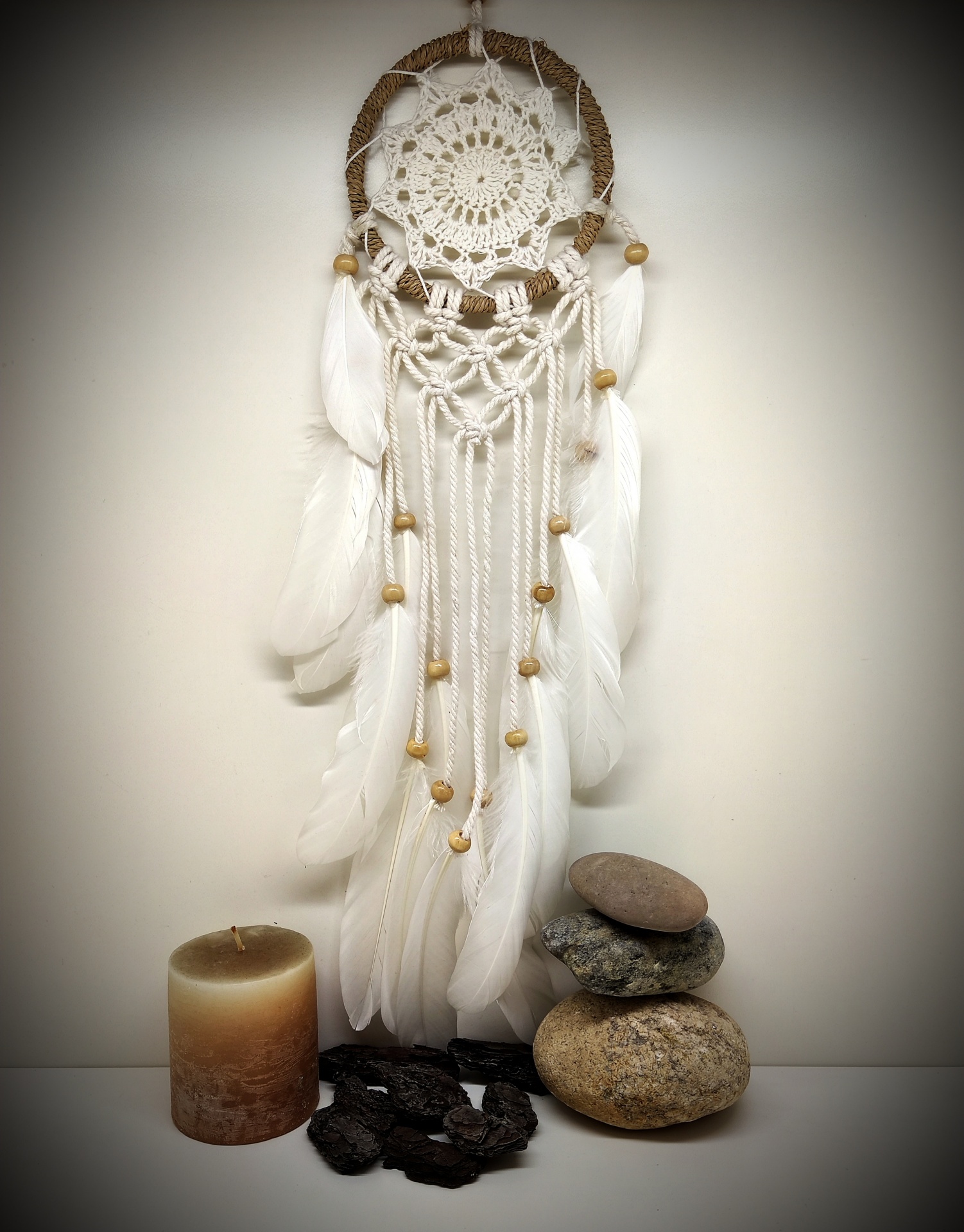 Apanha-sonhos de madeira com penas brancas e contas, vela castanha e pedras empilhadas ao lado