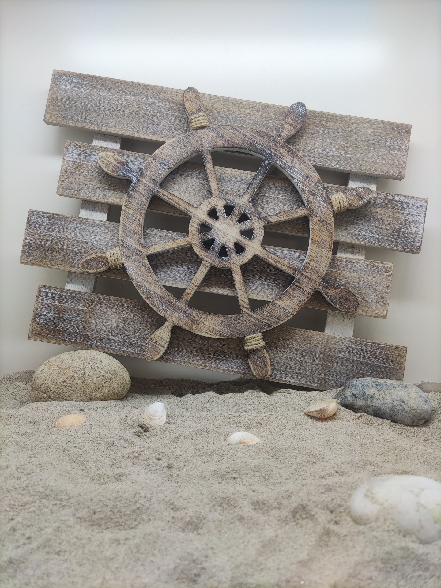 Roda de leme decorativa de madeira rústica sobre painel e areia com conchas e pedras