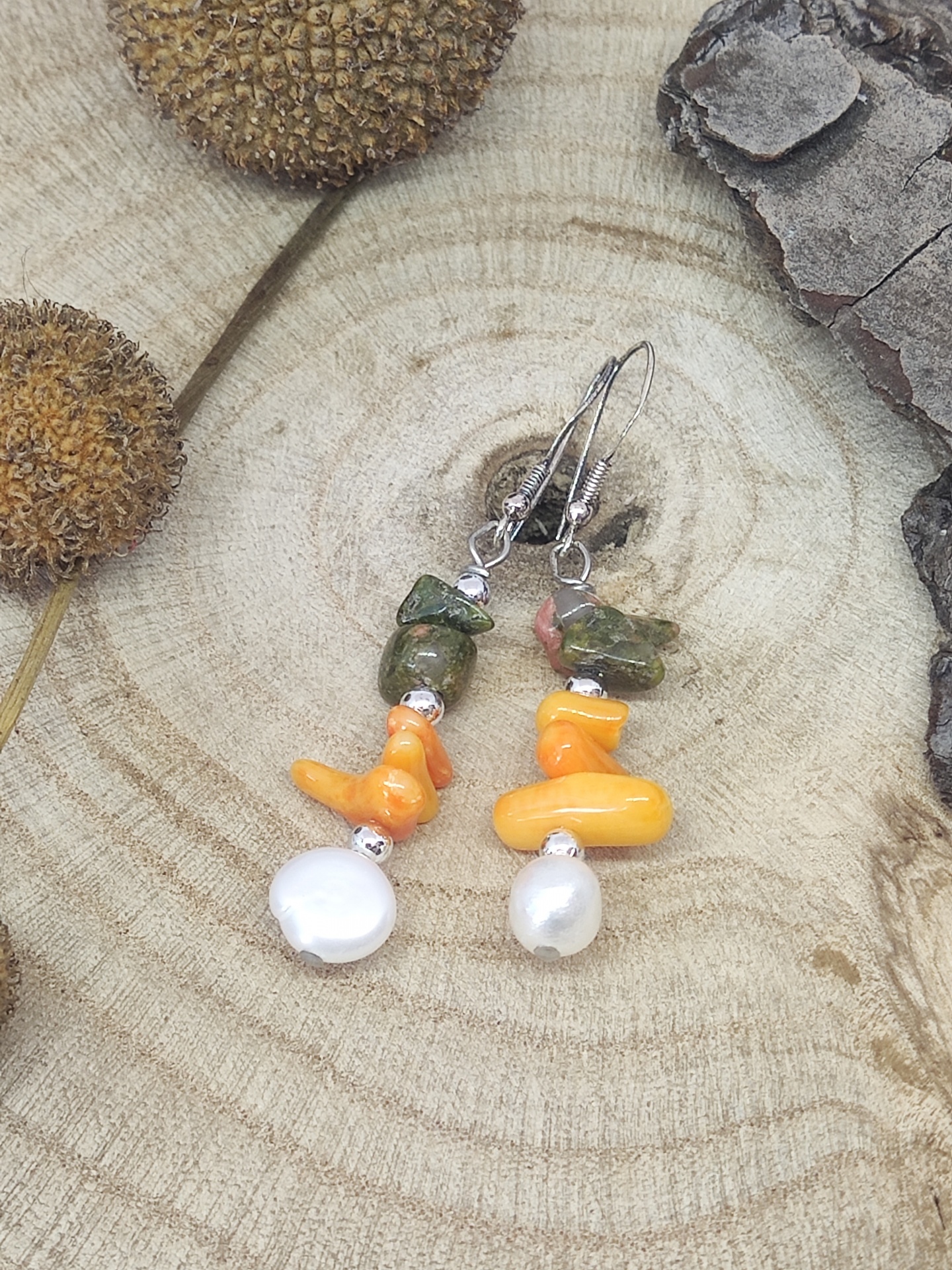 Brincos com contas verdes, laranja e pérolas sobre madeira com elementos naturais secos.