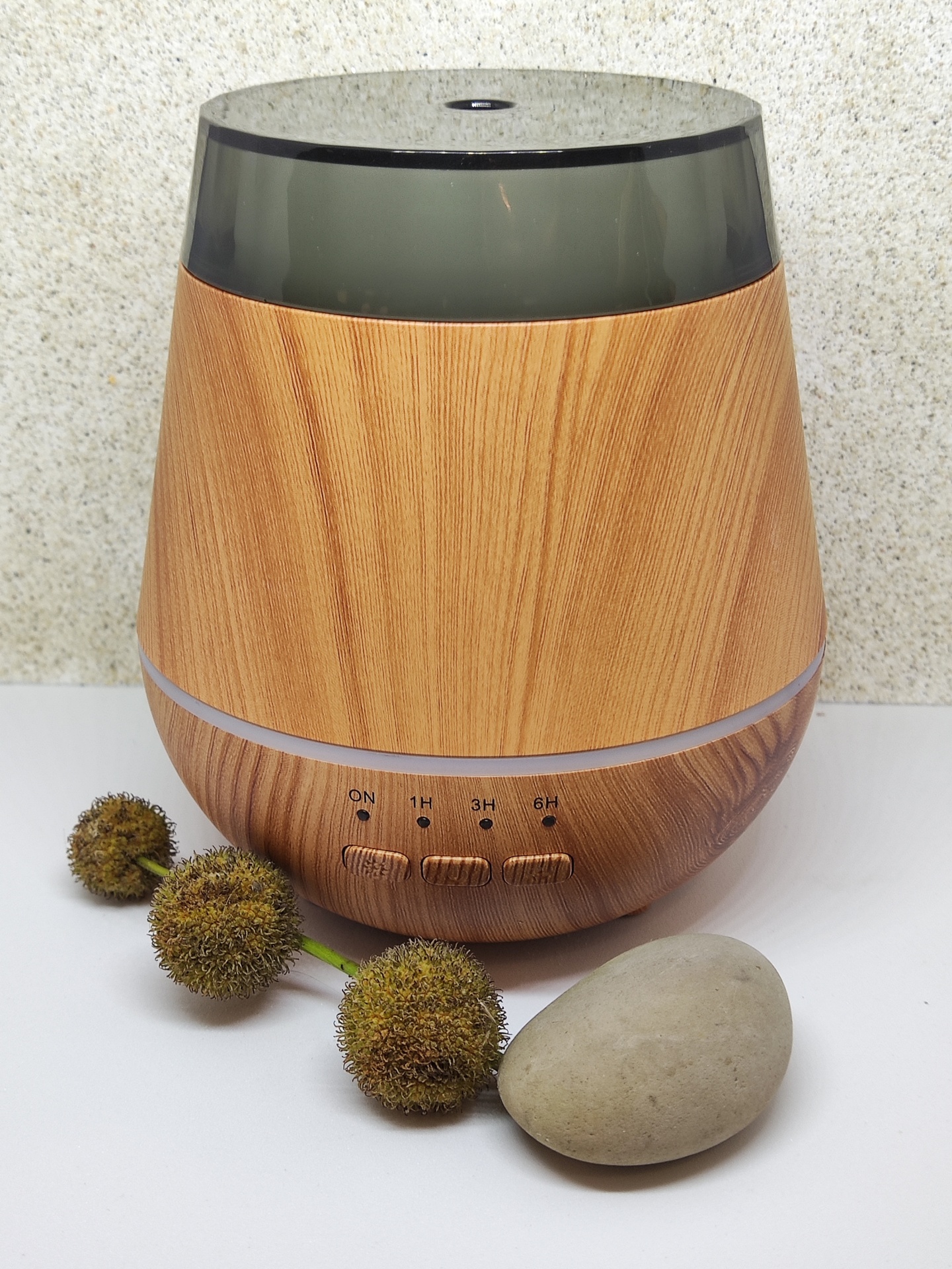 Difusor de aroma em madeira clara com cúpula translúcida escura e botões de controlo, com flores secas e pedra ao lado