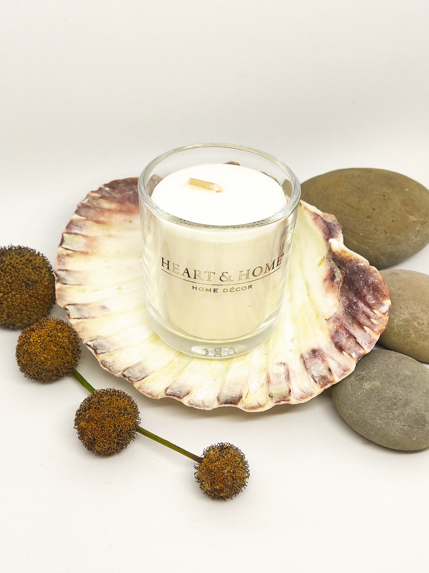 Vela em copo de vidro numa concha decorativa cercada de pedras e flores secas.