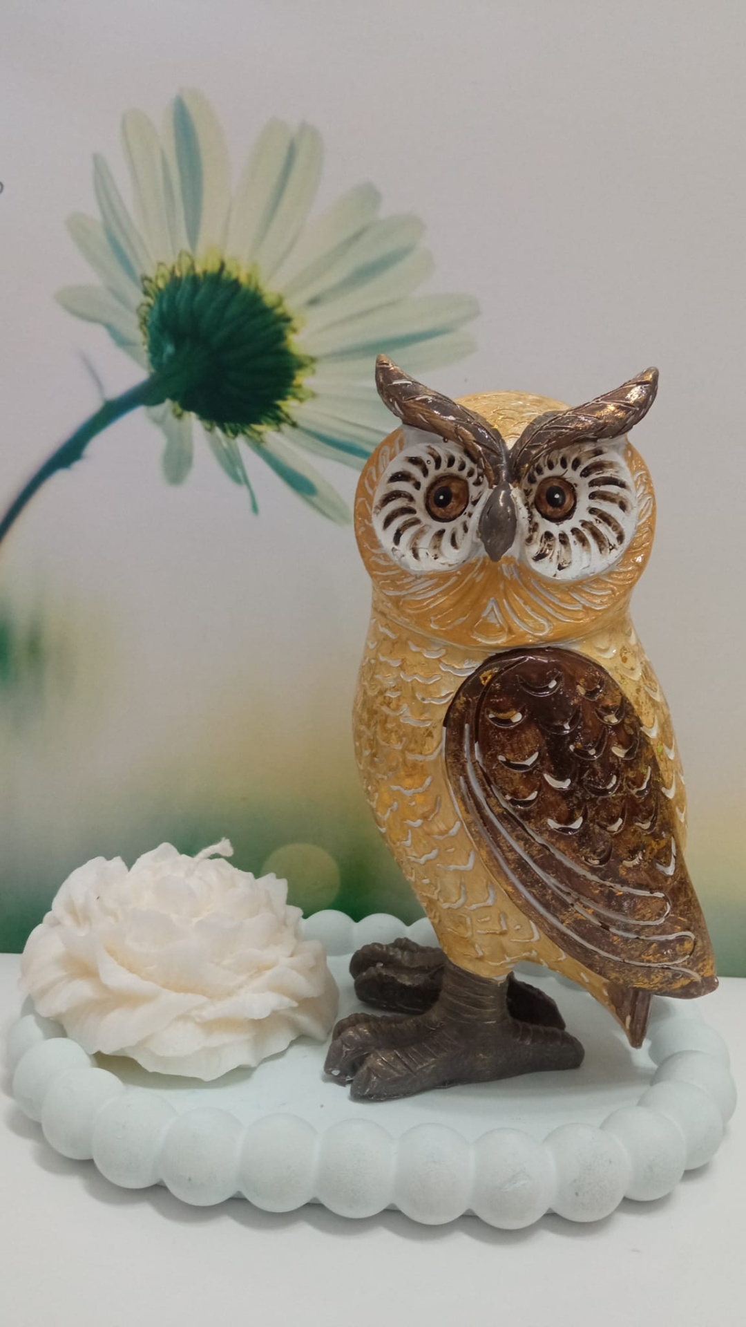 Estátua de coruja decorativa sobre base branca com flor artificial e fundo com flor margarida