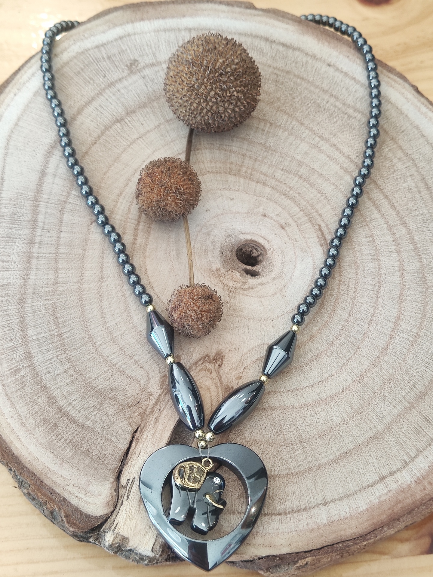 Colar cinza chumbo com pendente coração e emblema dourado sobre base de madeira