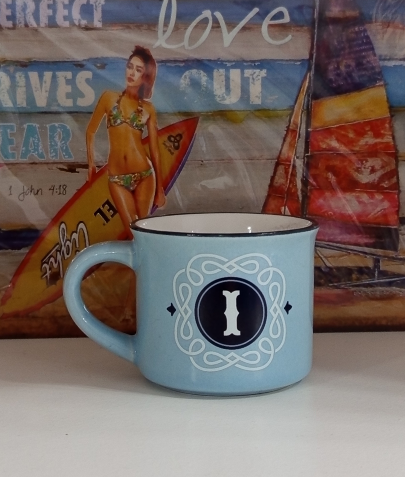 Caneca azul clara com letra I e desenho branco, com imagem de mulher e prancha ao fundo.