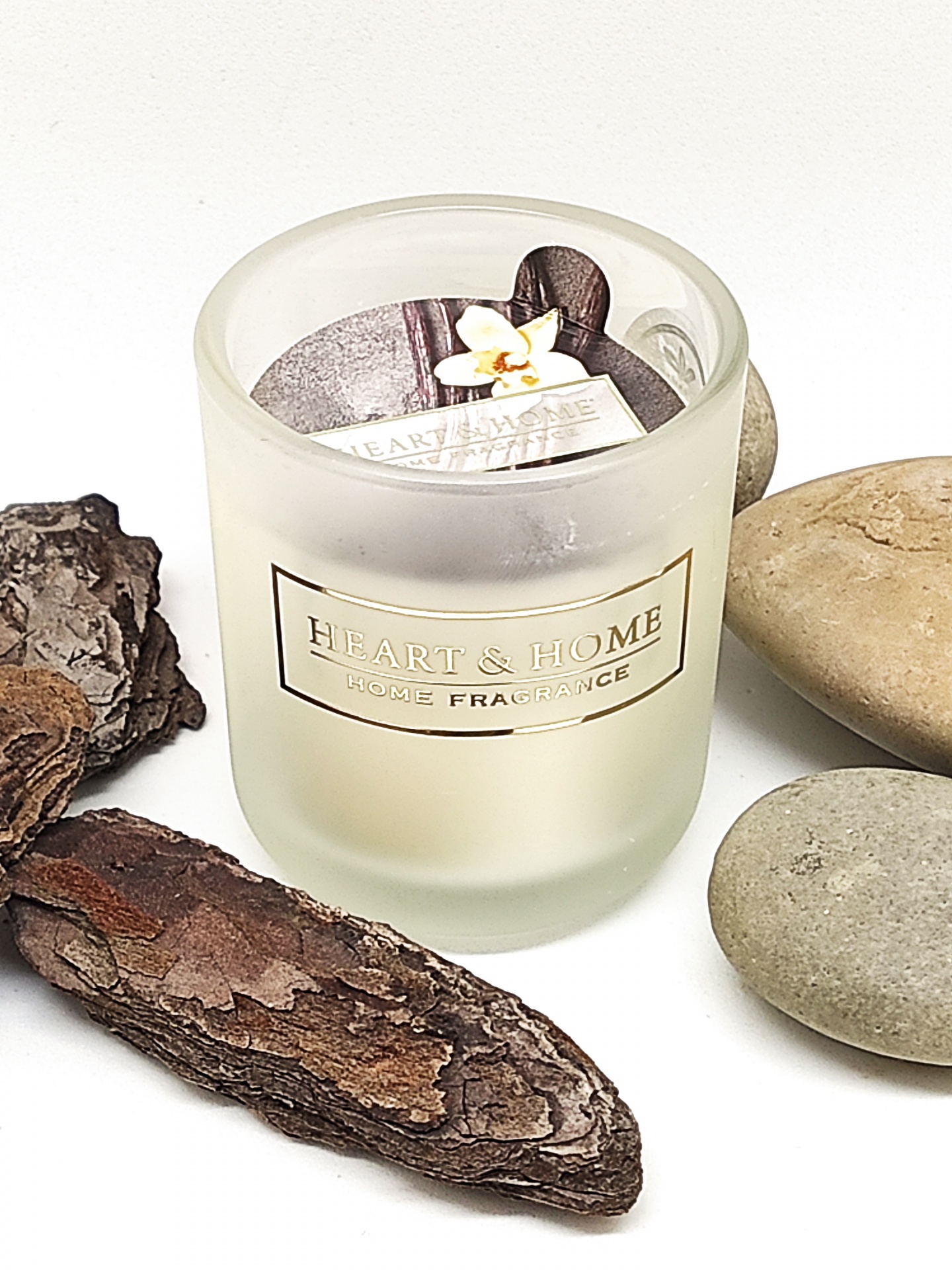 Vela aromática em copo de vidro fosco com etiqueta dourada ao centro, rodeada por casca de madeira e pedras lisas