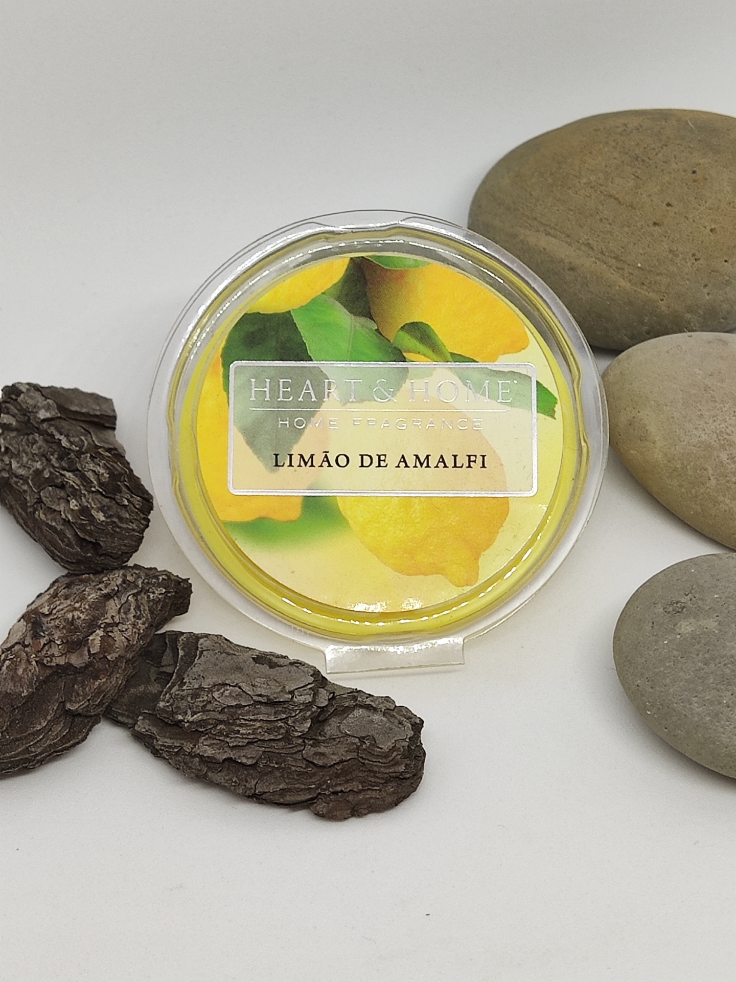 Produto redondo plástico com etiqueta de limões e texto, rodeado de cascas de árvore e pedras lisas