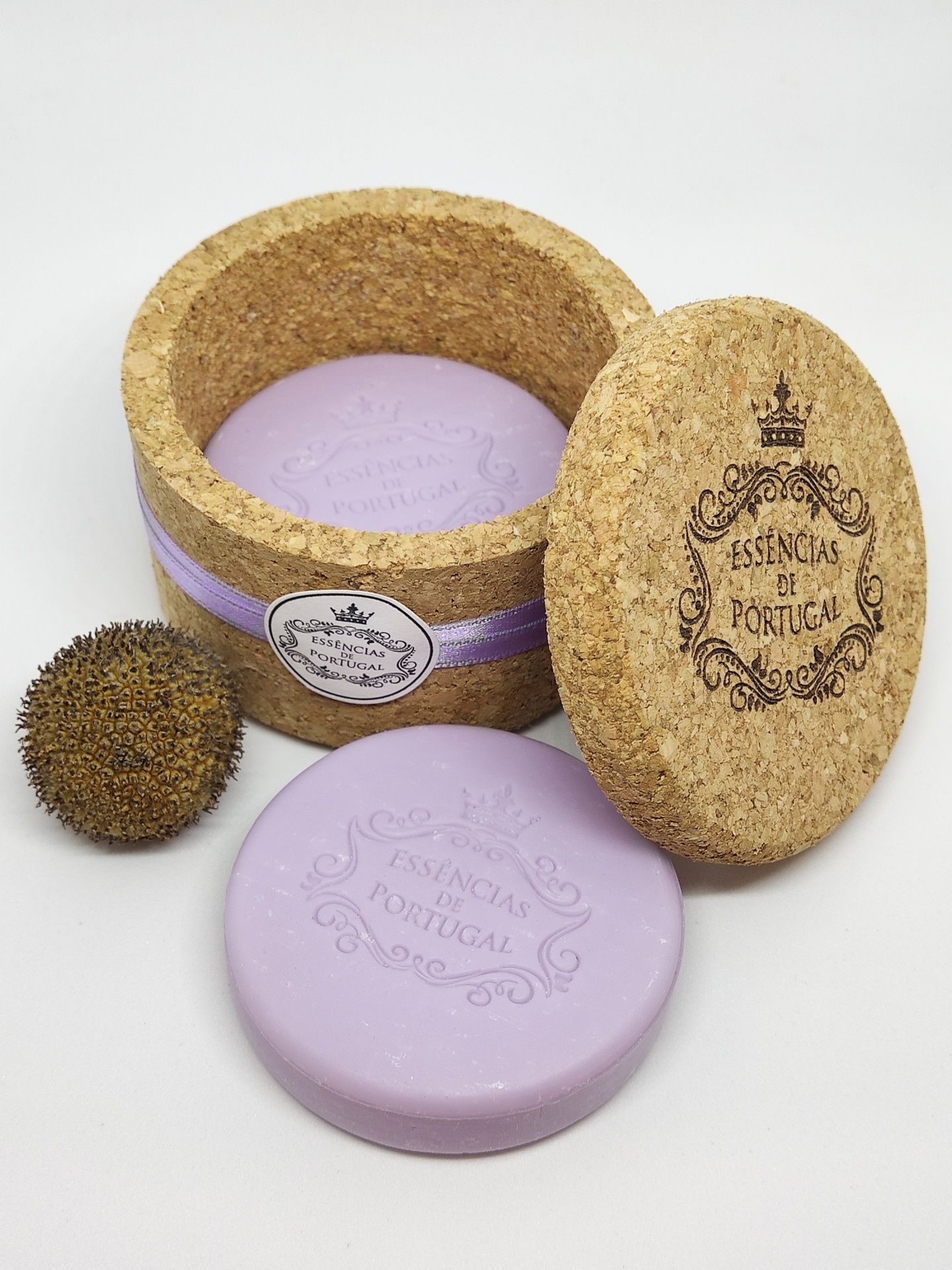 Sabonete lilás redondo dentro de caixa de cortiça redonda, com tampa gravada e fruto seco ao lado.