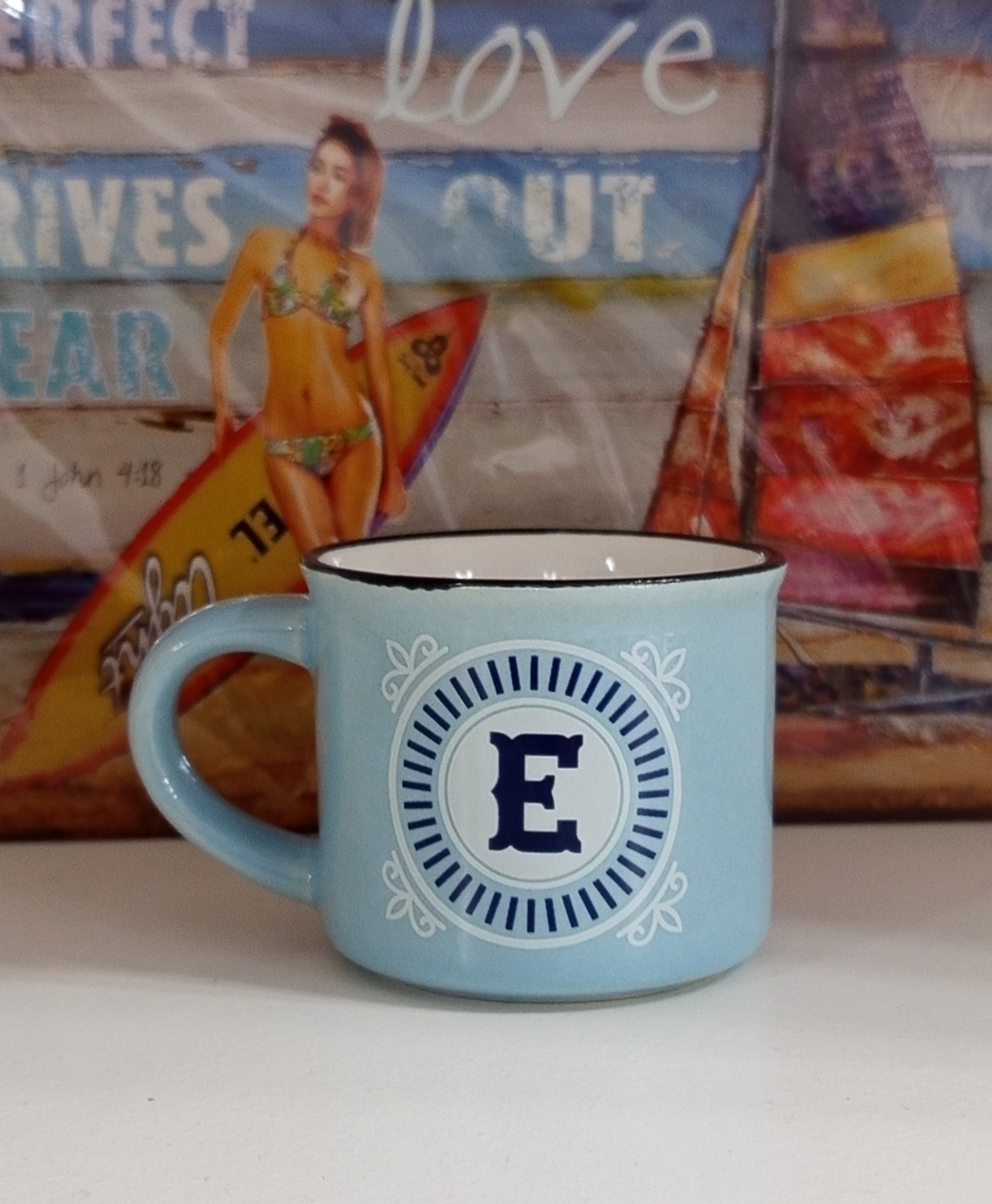 Caneca azul claro com a letra E decorativa, sobre superfície branca e fundo colorido.