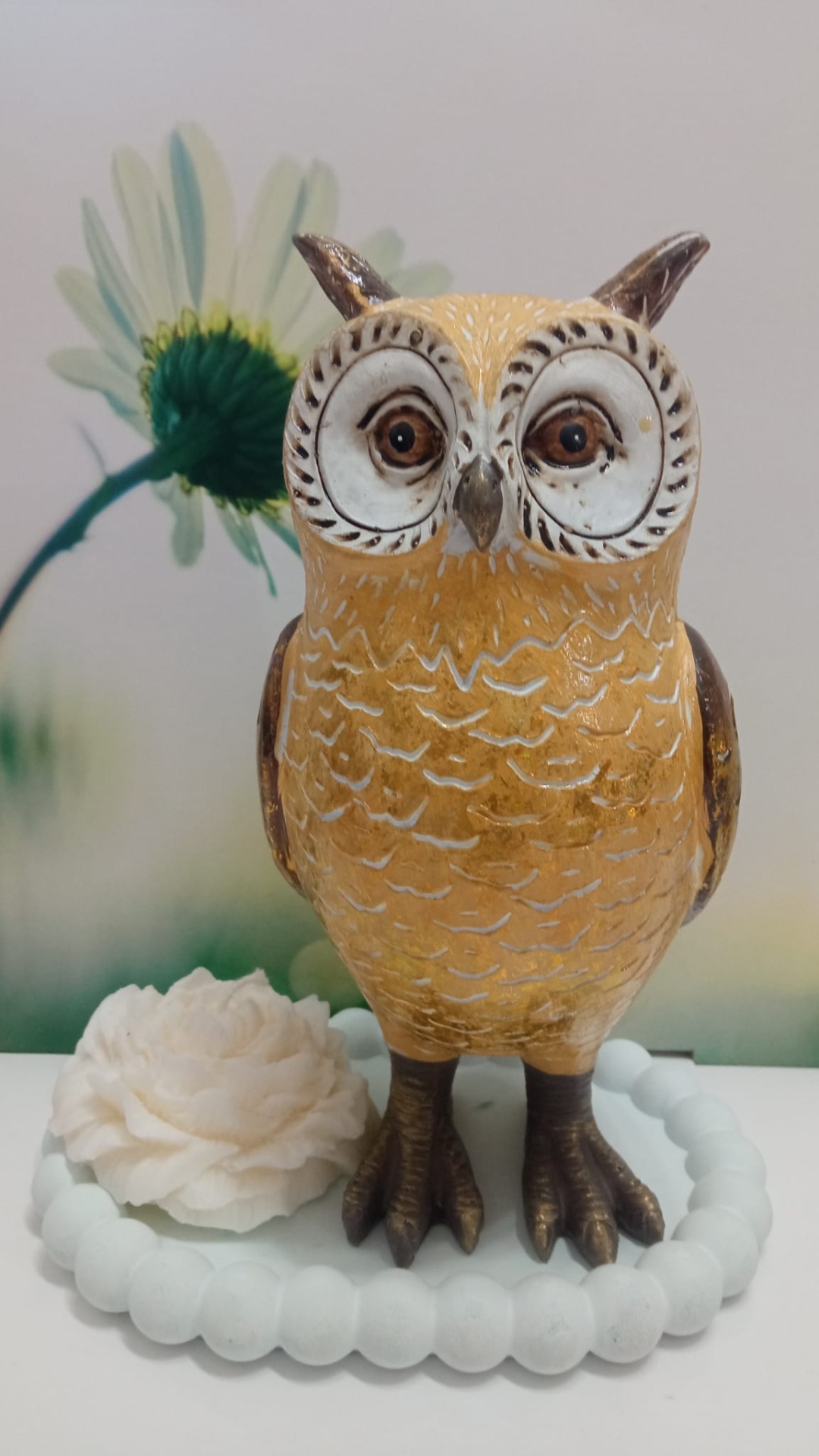 Estatueta de coruja castanha com olhos grandes ao lado de flor branca e pulseira de contas brancas