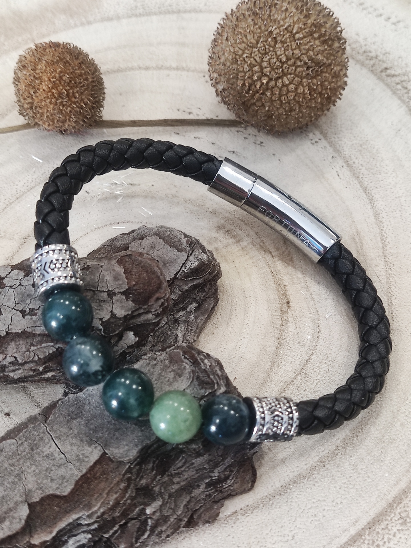 Pulseira preta trançada com contas verdes e fecho metálico sobre madeira clara