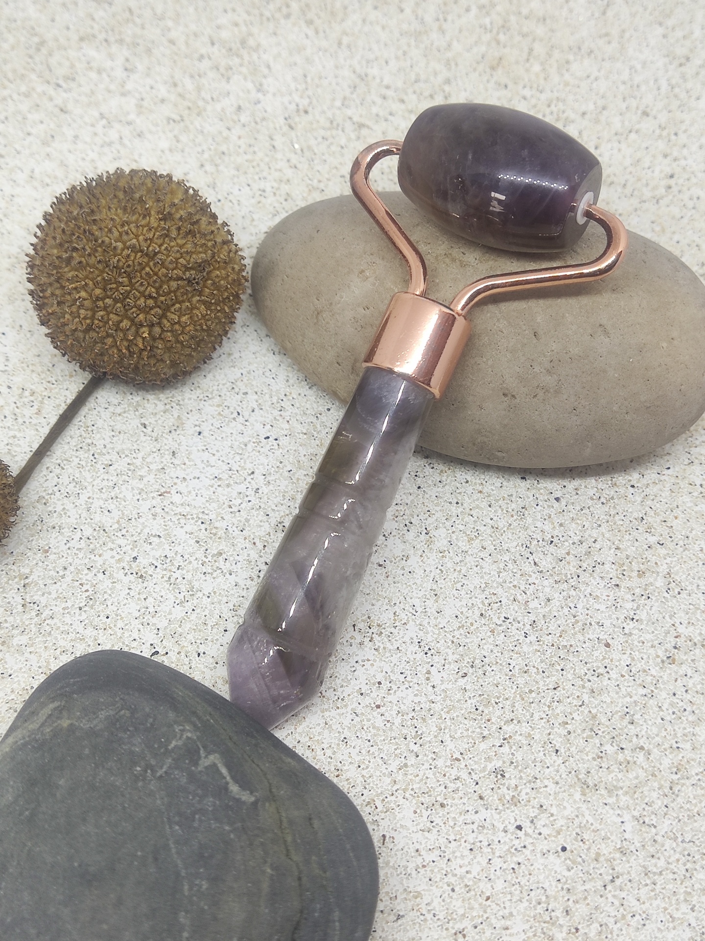 Rolo facial de pedra ametista com suporte de metal cor cobre sobre pedras e planta seca