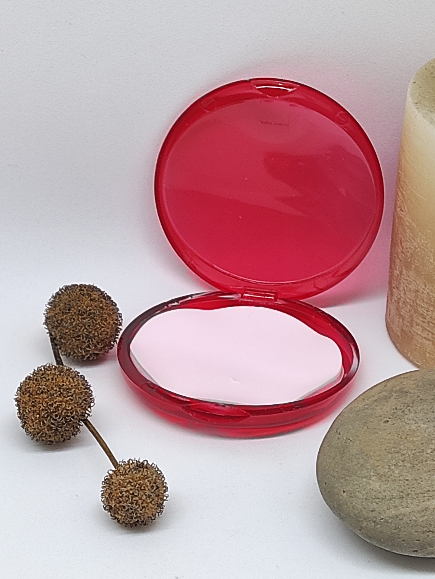 Compacto vermelho aberto com pó branco, bolbos secos, vela e pedra sobre fundo branco.