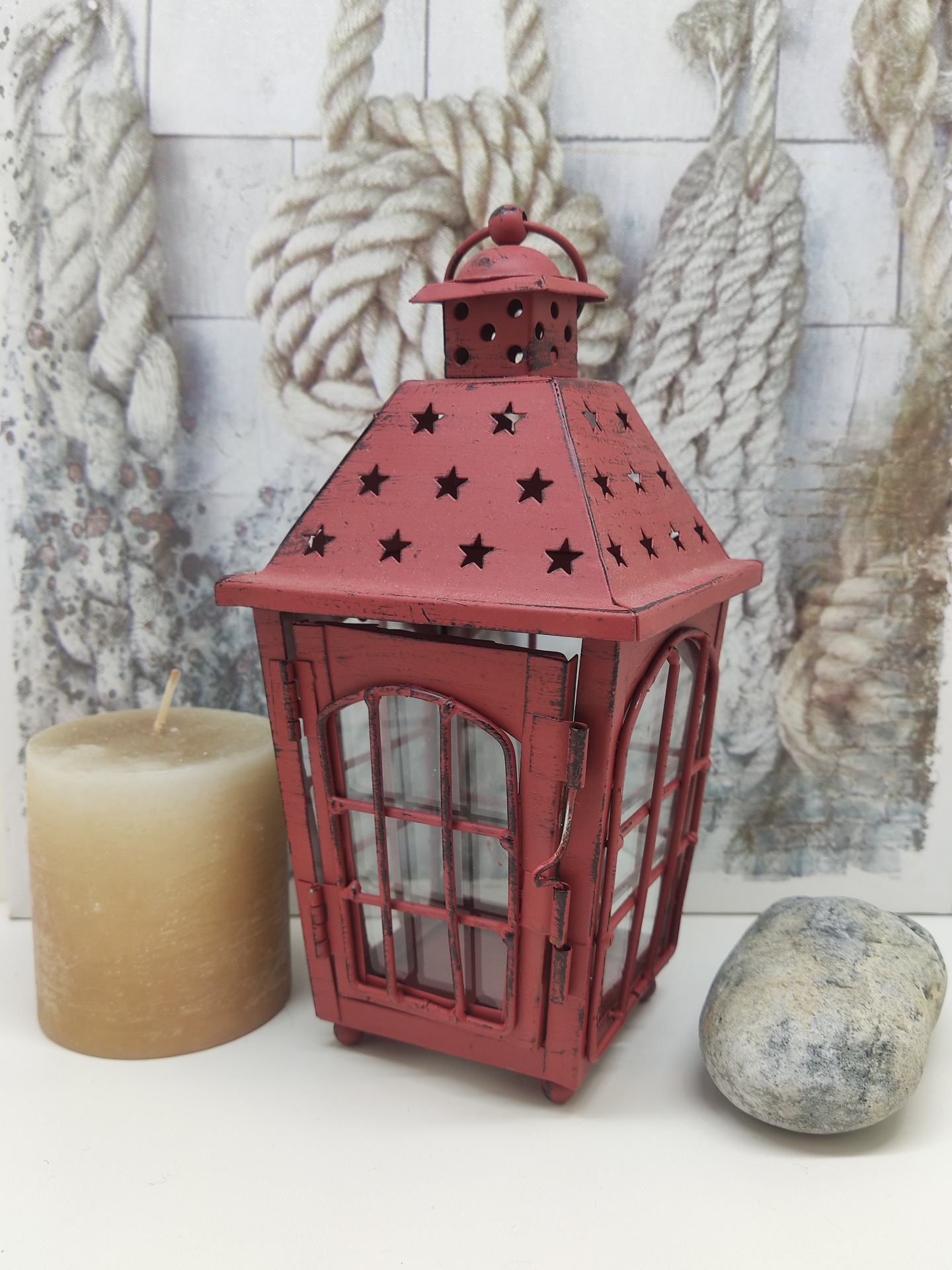 Lanterna decorativa vermelha metálica com estrelas, vela bege e pedra cinzenta em fundo com cordas