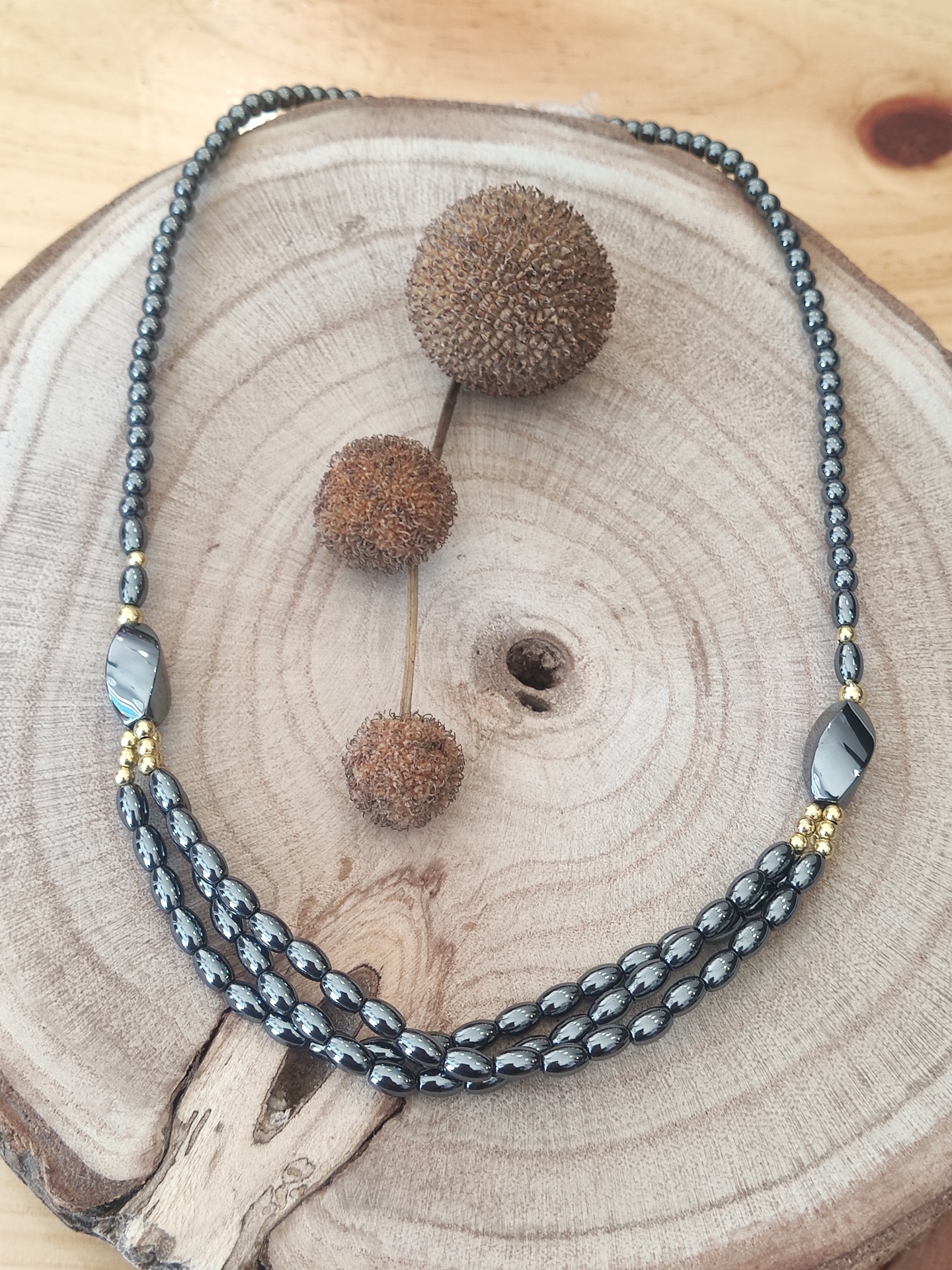 Colar com contas cinza e douradas sobre base de madeira redonda com sementes secas