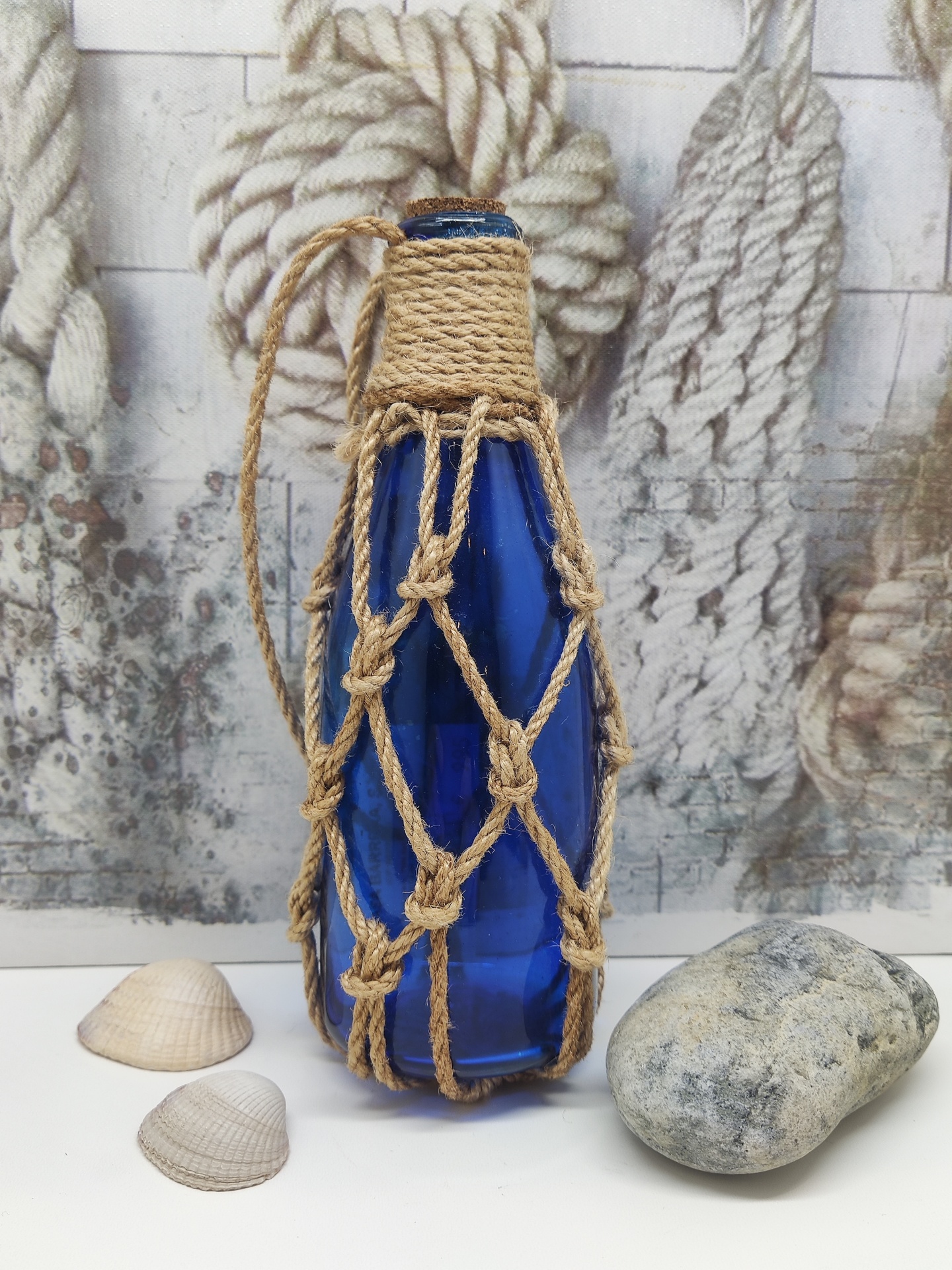 Garrafa azul de vidro com suporte de corda em padrão de rede, moluscos e pedra ao lado