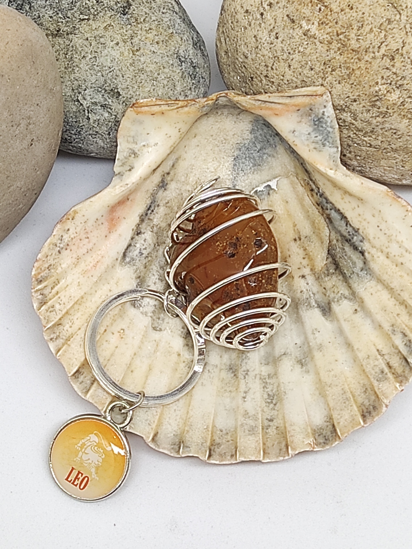 Chaveiro com pedra castanha em fio de metal e pendente amarelo com texto LEO, numa concha, pedras ao fundo