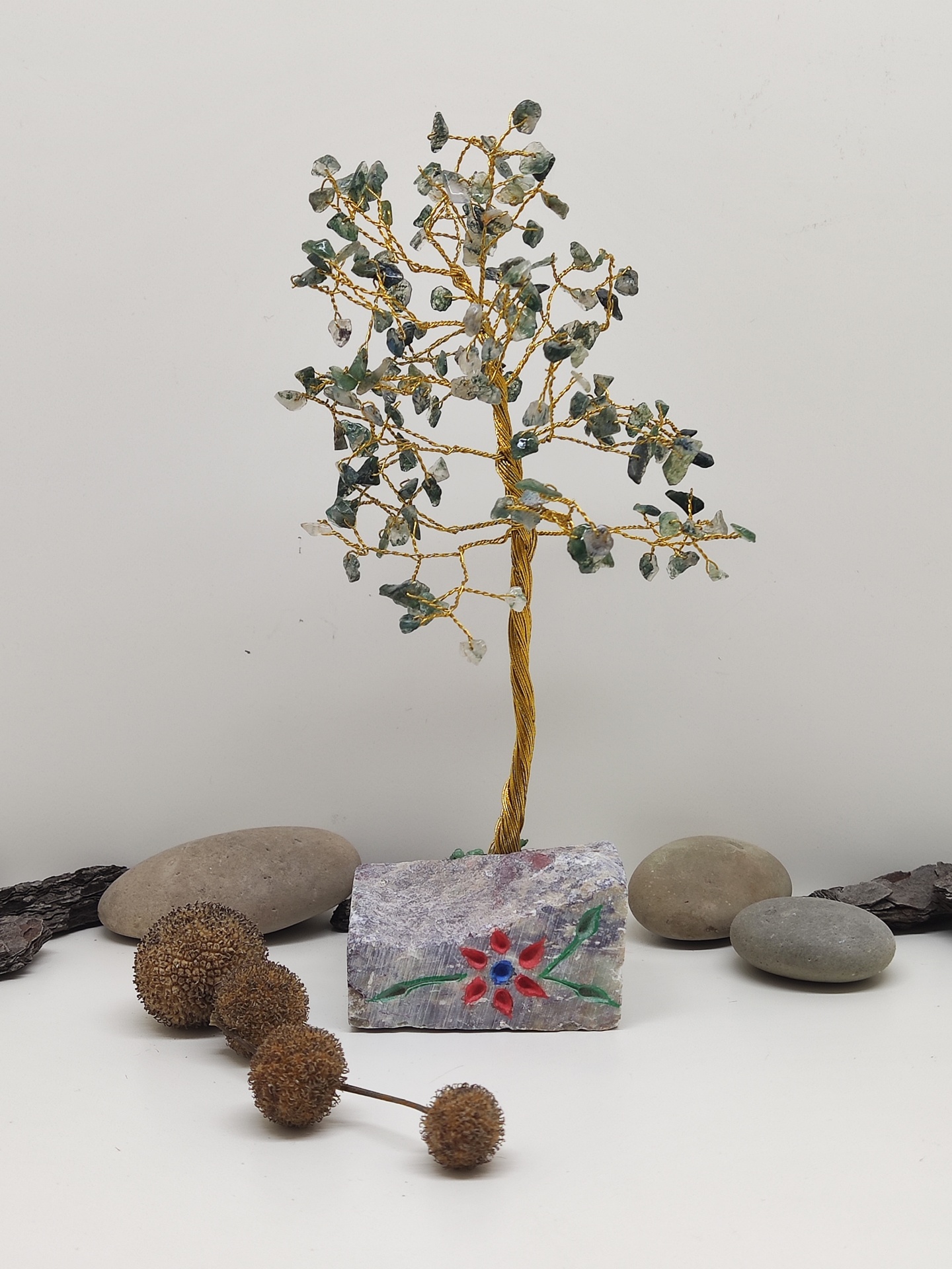 Árvore decorativa de fios dourados e pedras verdes numa base de pedra pintada com flor, rodeada por pedras naturais e ramos secos.