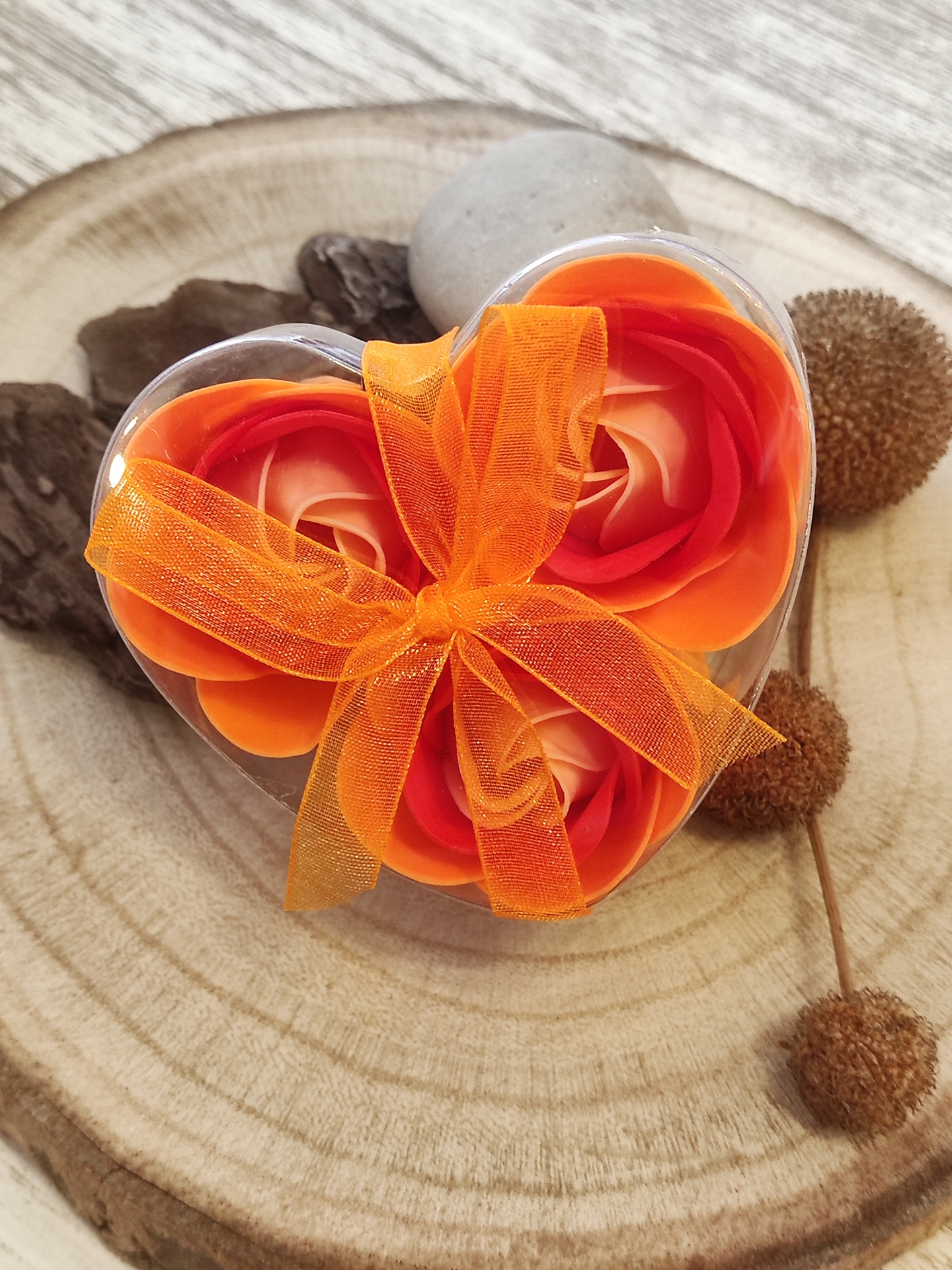 Caixa coração transparente com flores artificiais laranja e creme e laço de tule laranja sobre madeira