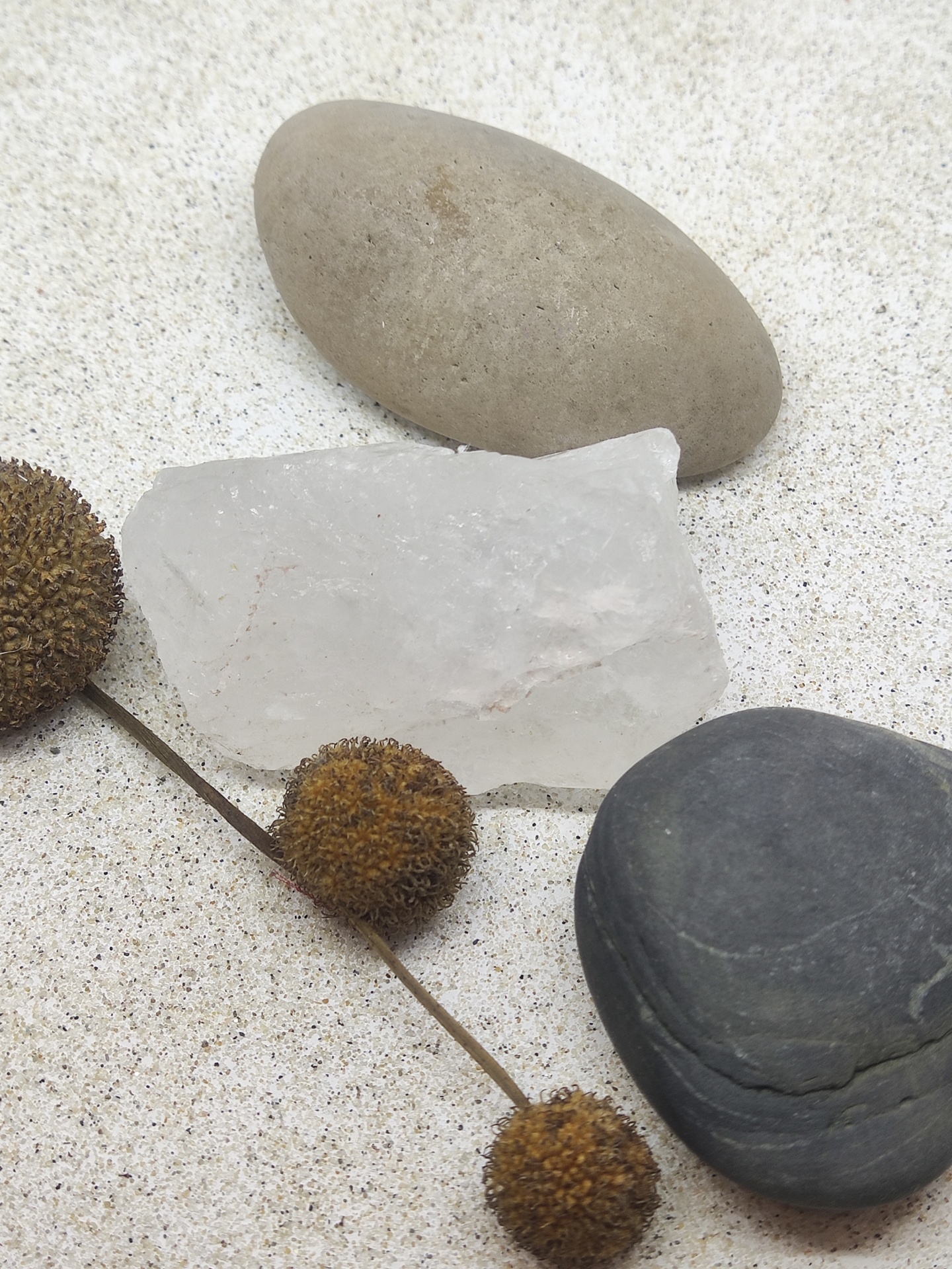 Três pedras lisas em tons bege, branco e preto com um ramo seco com bolas castanhas em fundo branco granuloso