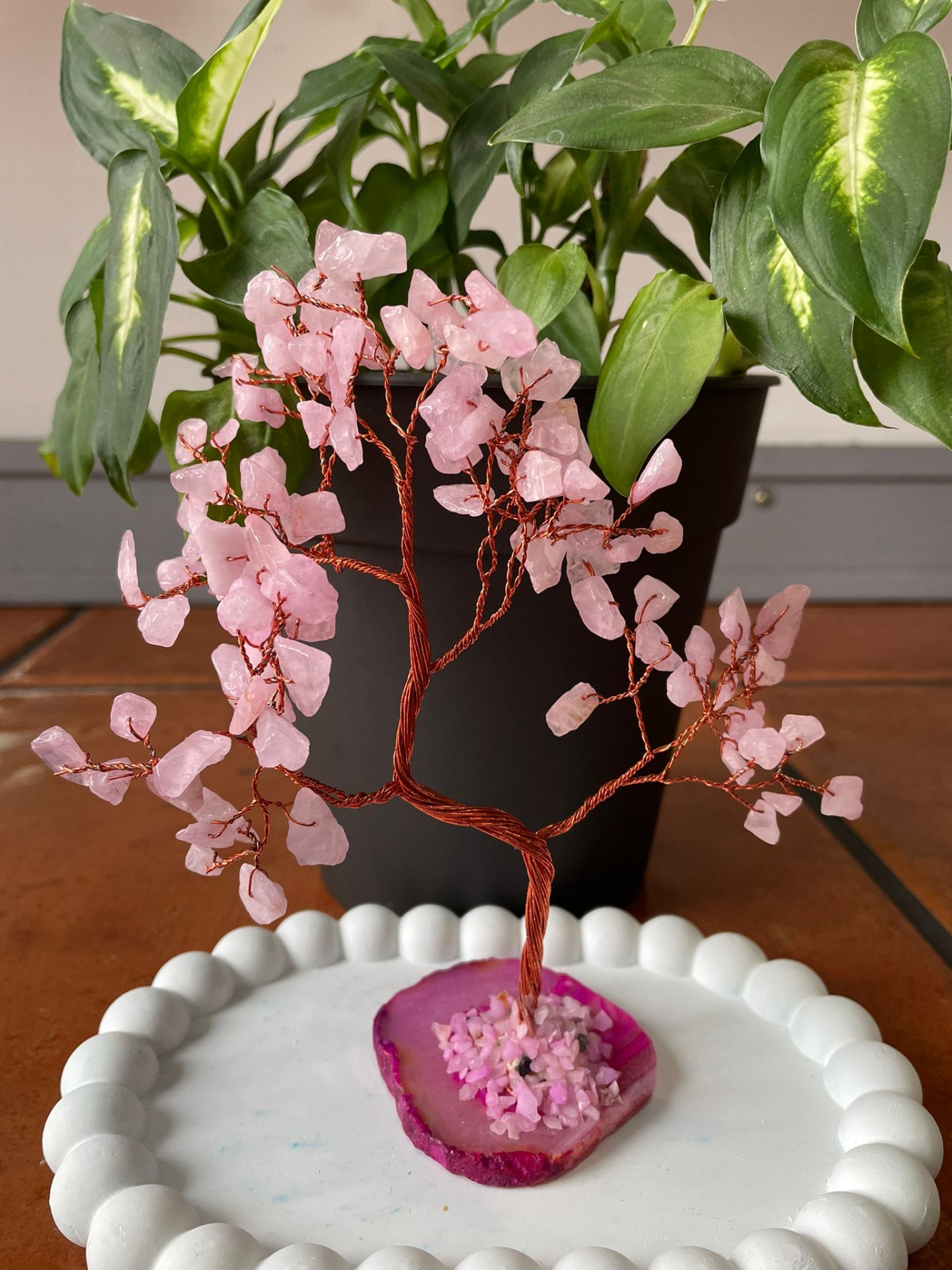 Árvore decorativa com pedras cor-de-rosa e base de ametista sobre superfície branca, planta verde em vaso preto ao fundo