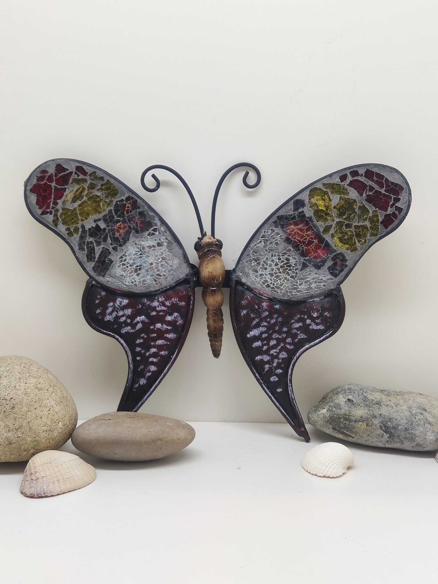 Escultura decorativa de borboleta com asas em mosaico colorido sobre superfície branca com pedras e conchas
