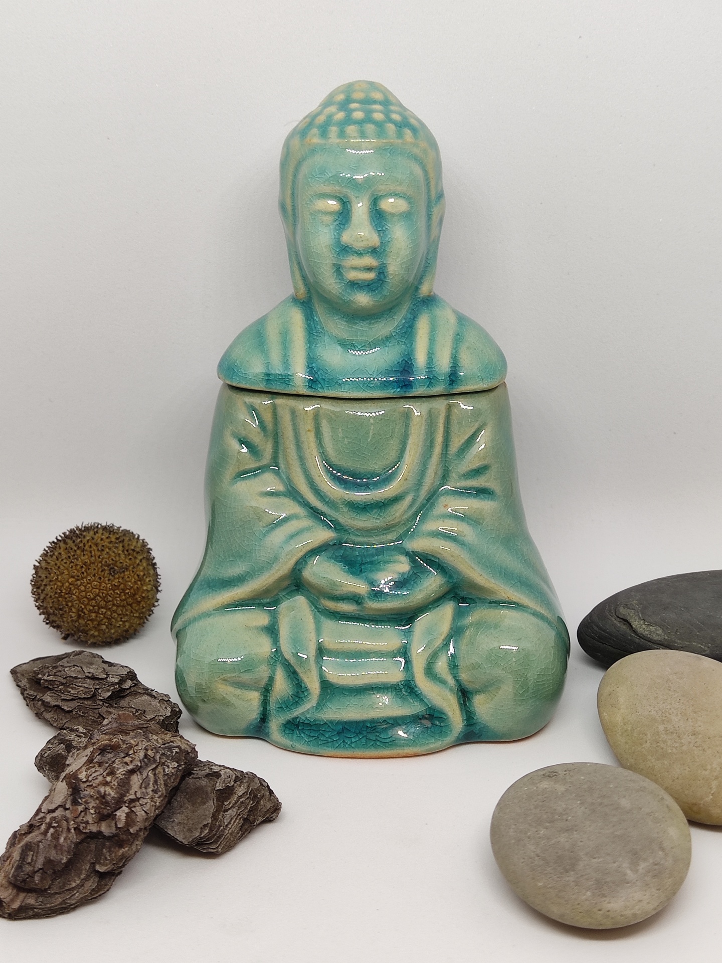 Frasco cerâmico azul-esverdeado em forma de Buda com pedras naturais em fundo branco.