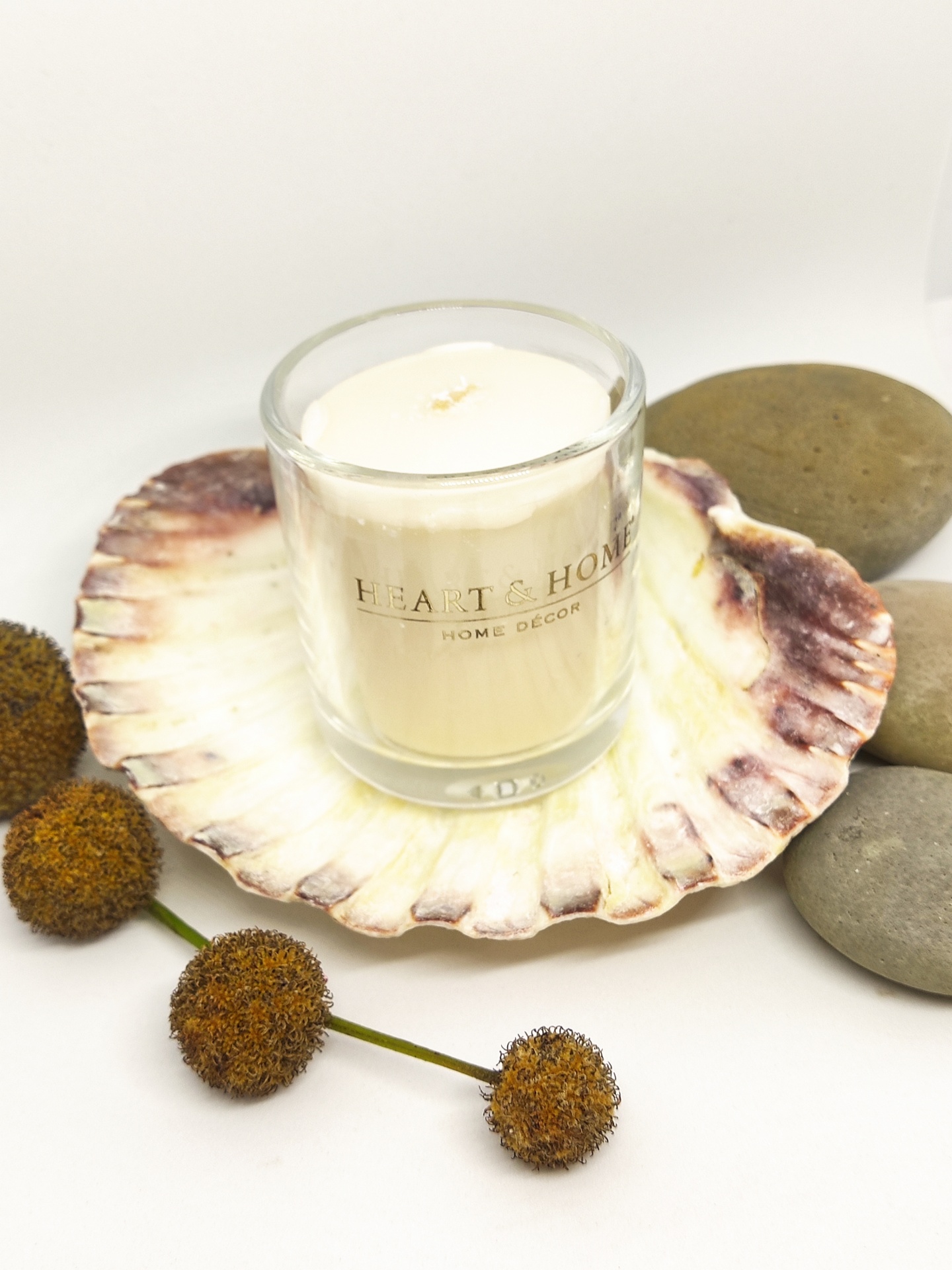 Vela branca em copo de vidro com texto dourado sobre concha natural, pedras lisas e bolas secas.