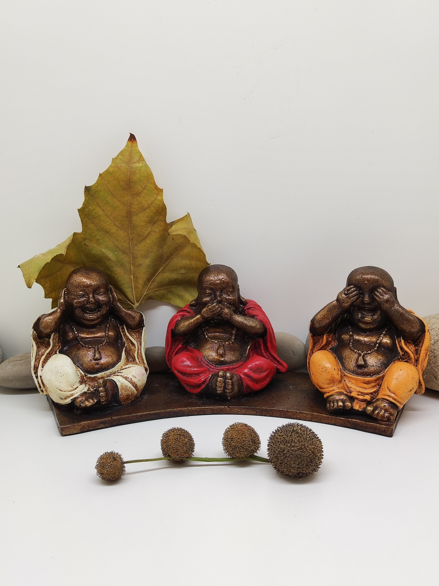 Três estatuetas Buda com roupas coloridas sobre base, folha seca atrás, frutos redondos na frente.