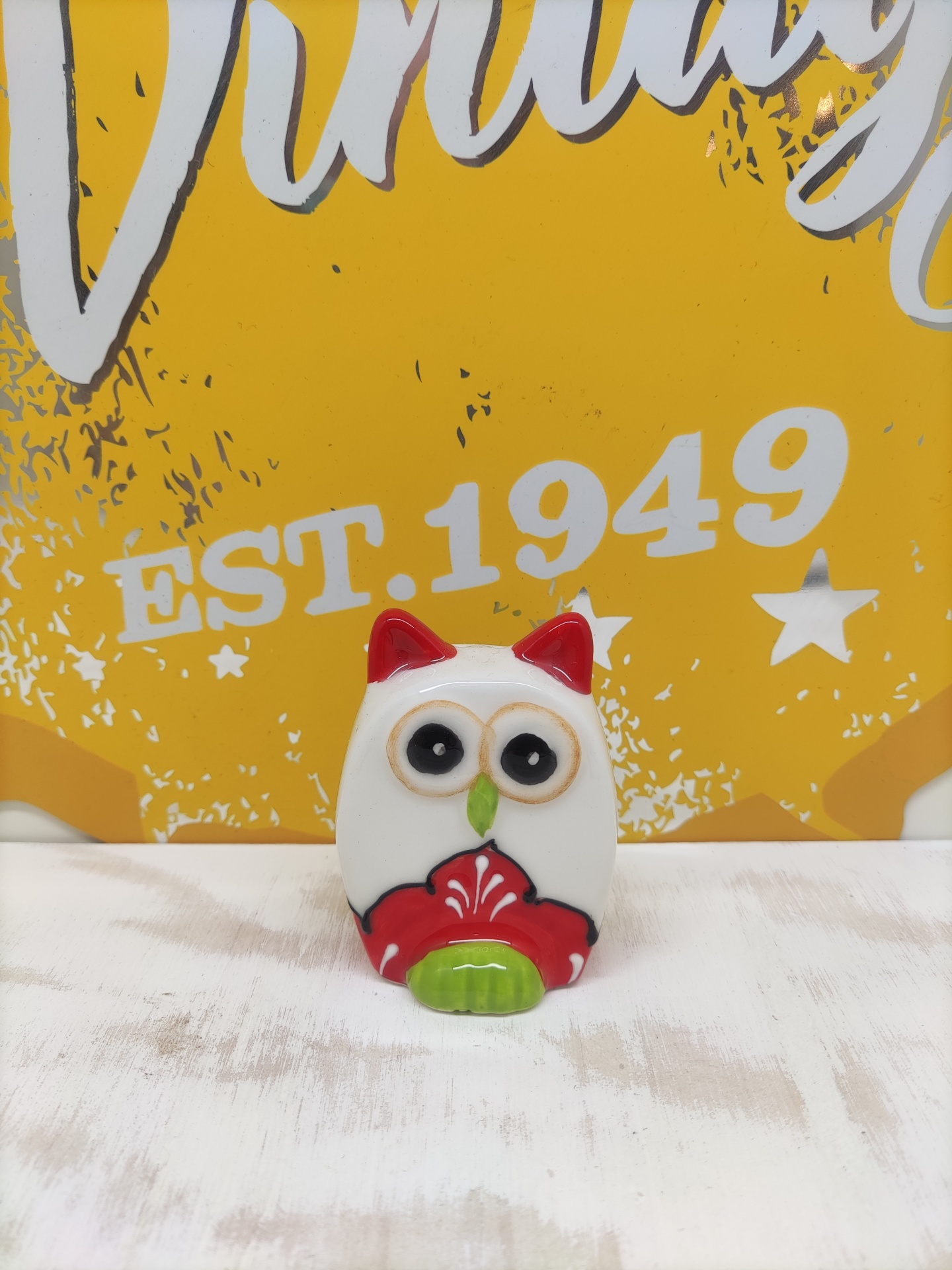 Figura decorativa de coruja em cerâmica com detalhes vermelhos e verdes diante de fundo amarelo com texto EST. 1949