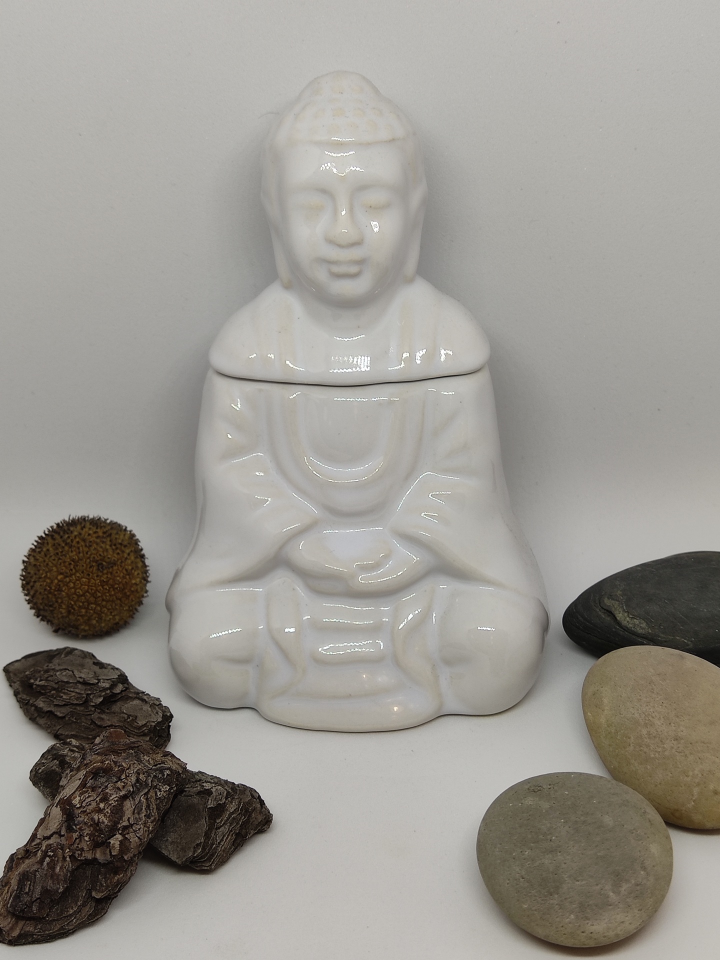 Escultura branca de figura meditativa com pedras e elemento vegetal seco ao redor.