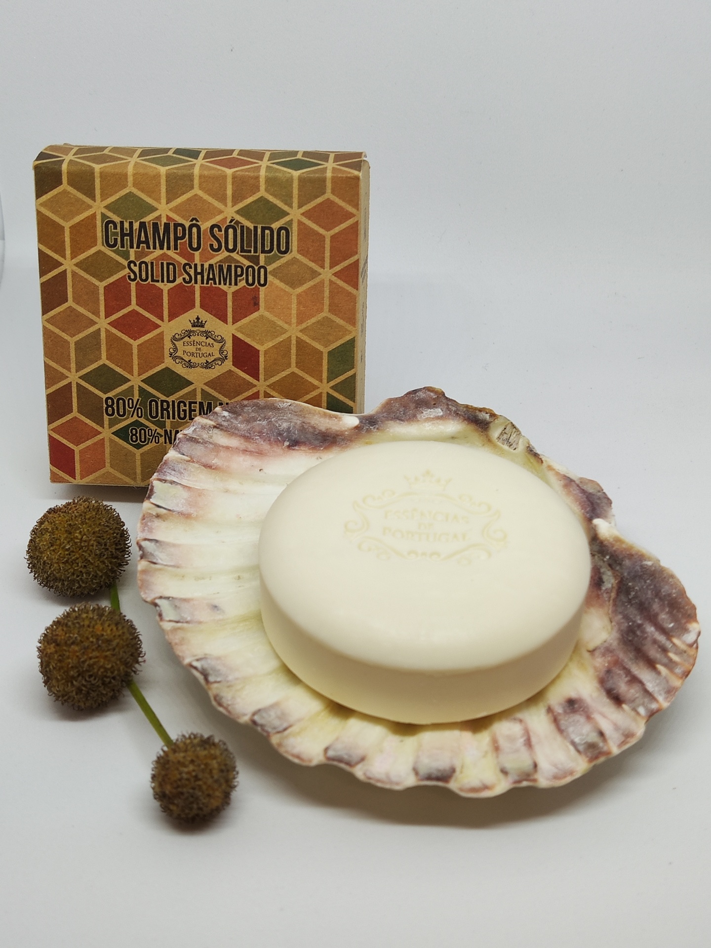 Champô sólido creme numa concha com embalagem geométrica atrás e flores castanhas ao lado