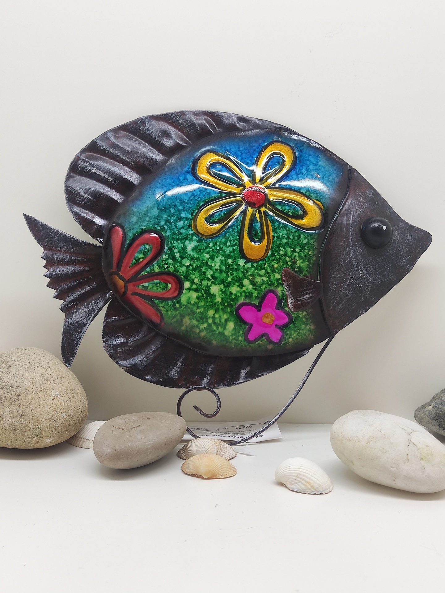 Peixe decorativo em metal colorido com flores, rodeado de pedras e conchas
