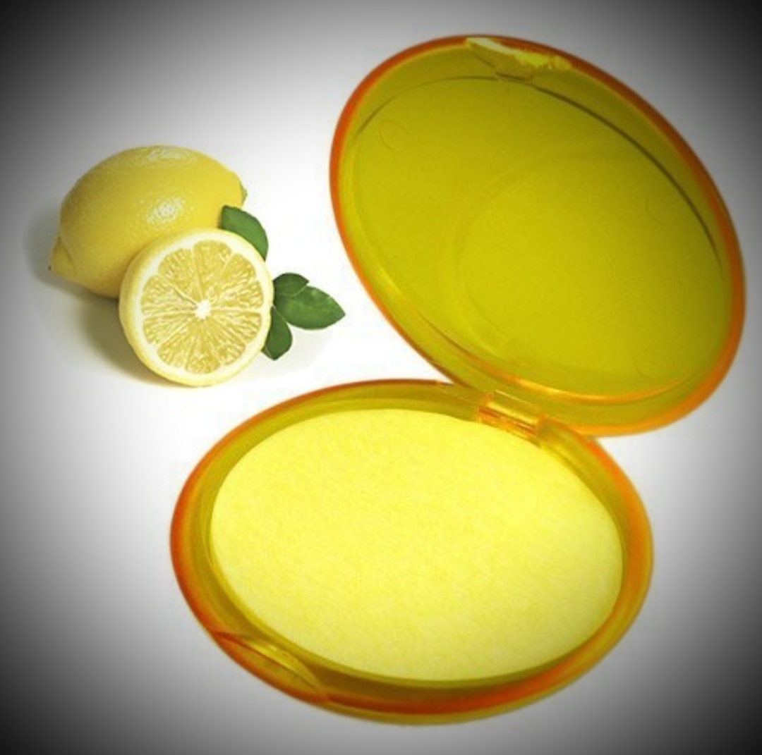 Pó compacto amarelo aberto ao lado de limões com folhas verdes