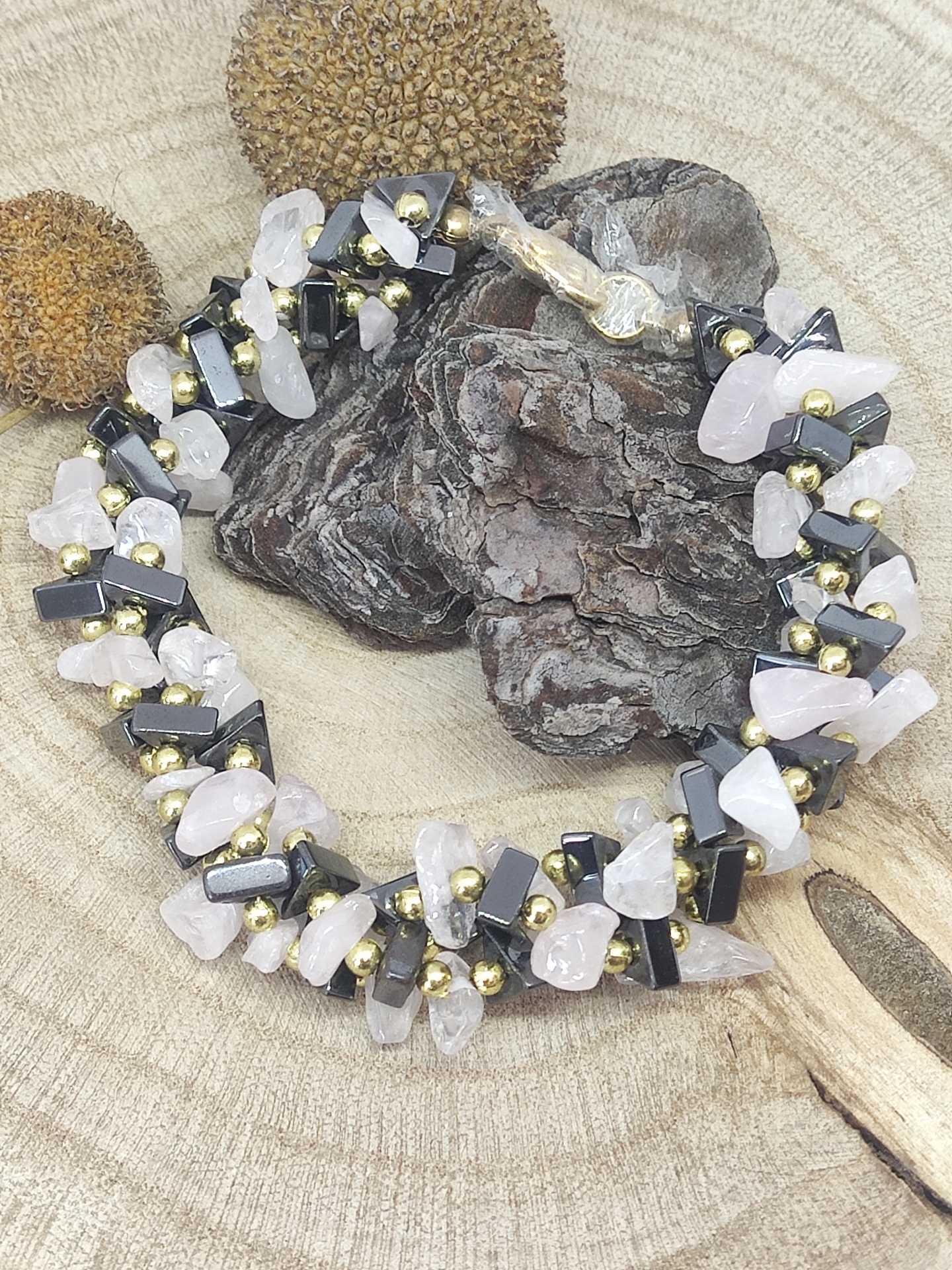 Colar de pedras rosa claro, pretas e esferas douradas sobre tronco de madeira com sementes e pedaço de madeira