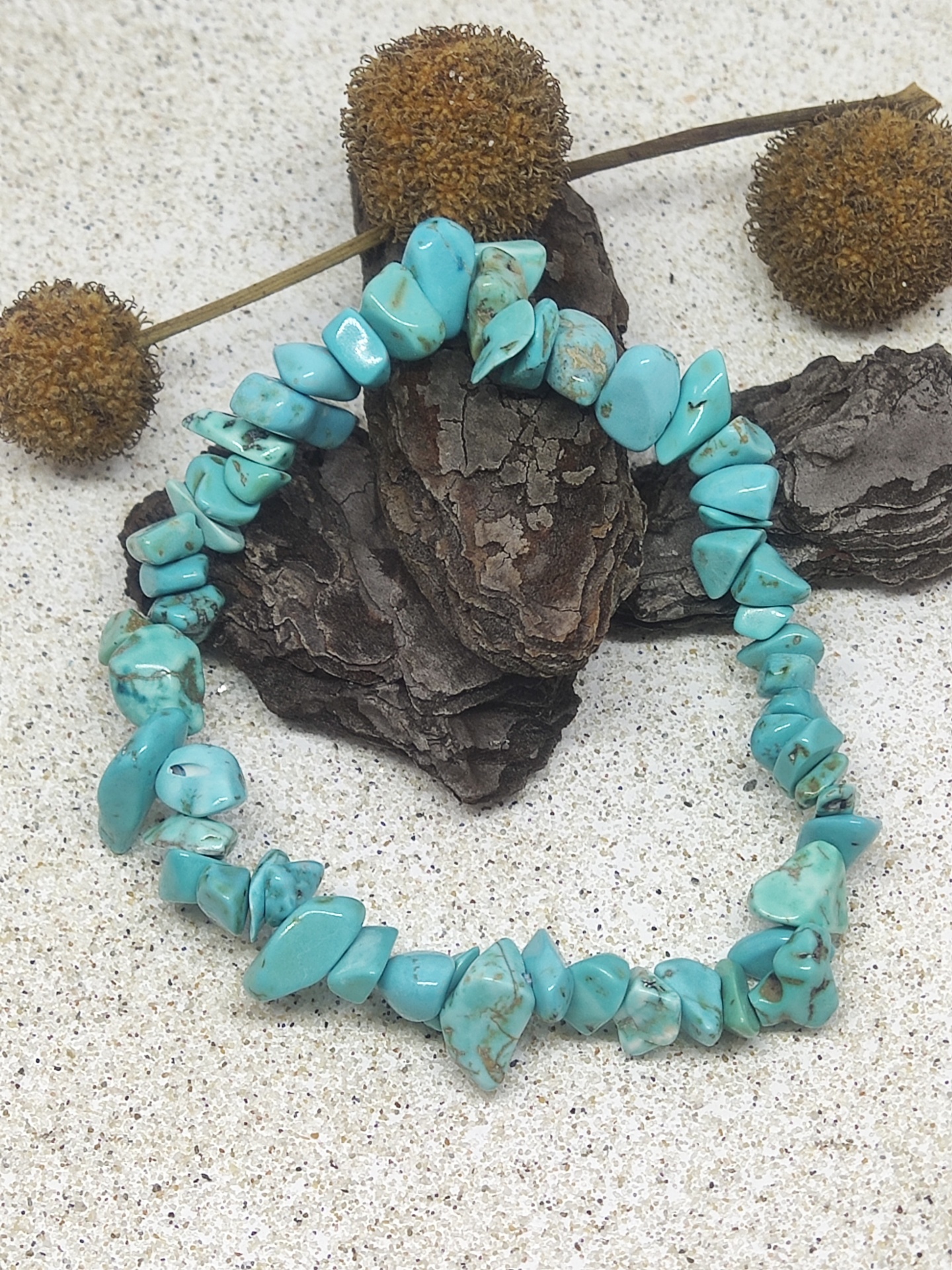 Pulseira de pedras azuis irregulares com manchas castanhas em fundo de areia e cascas de árvore