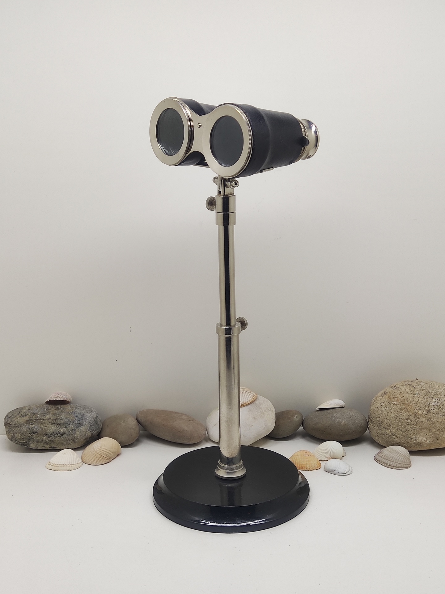 Binóculos antigos em suporte metálico com base preta, rodeados por pedras e conchas.