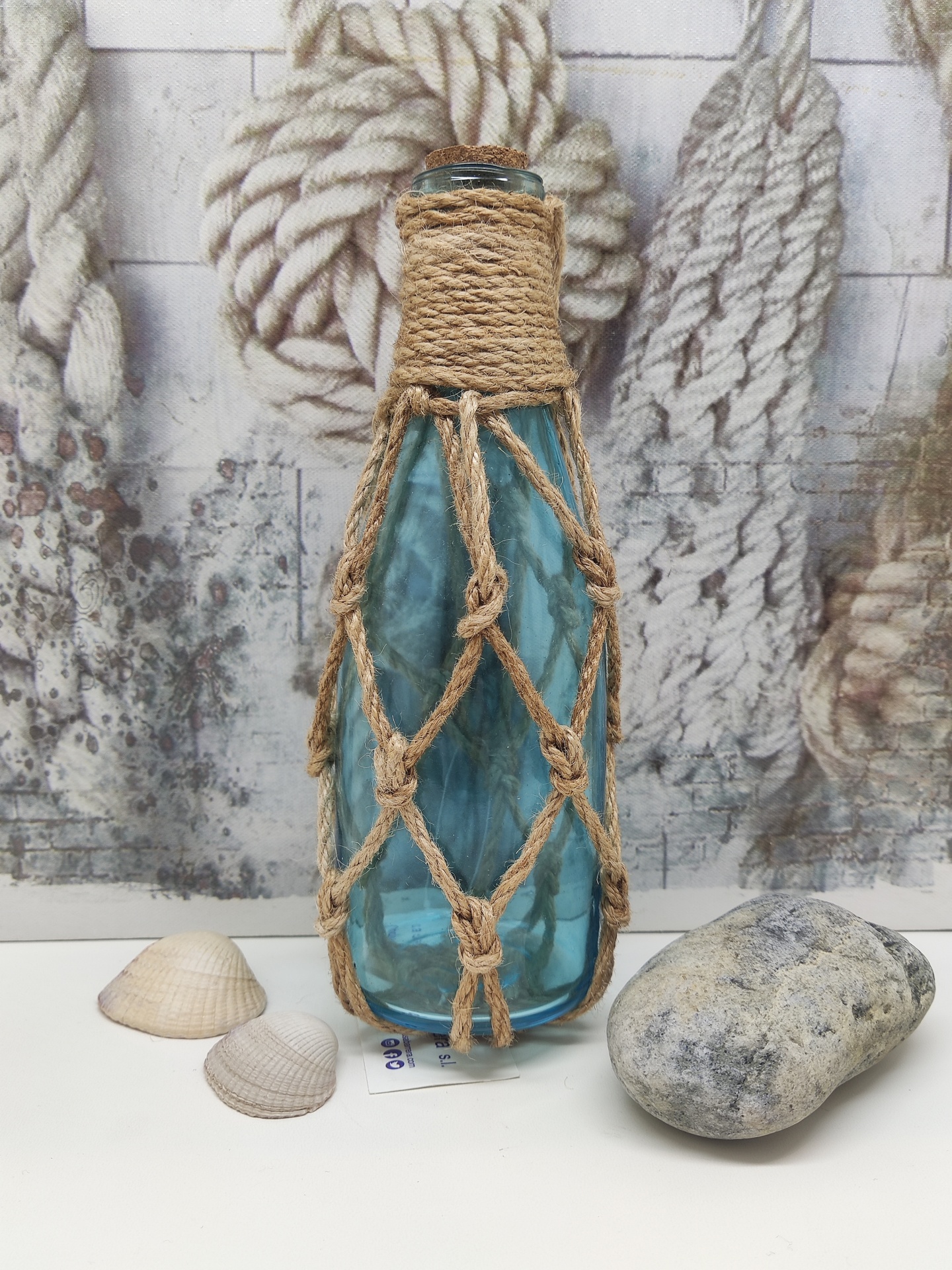 Garrafa de vidro azul com rede de corda natural, conchas e pedra sobre superfície branca