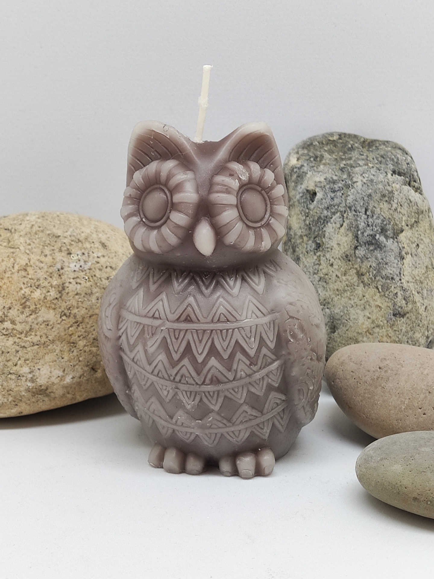 Vela castanha em forma de coruja com padrões geométricos, rodeada por pedras lisas