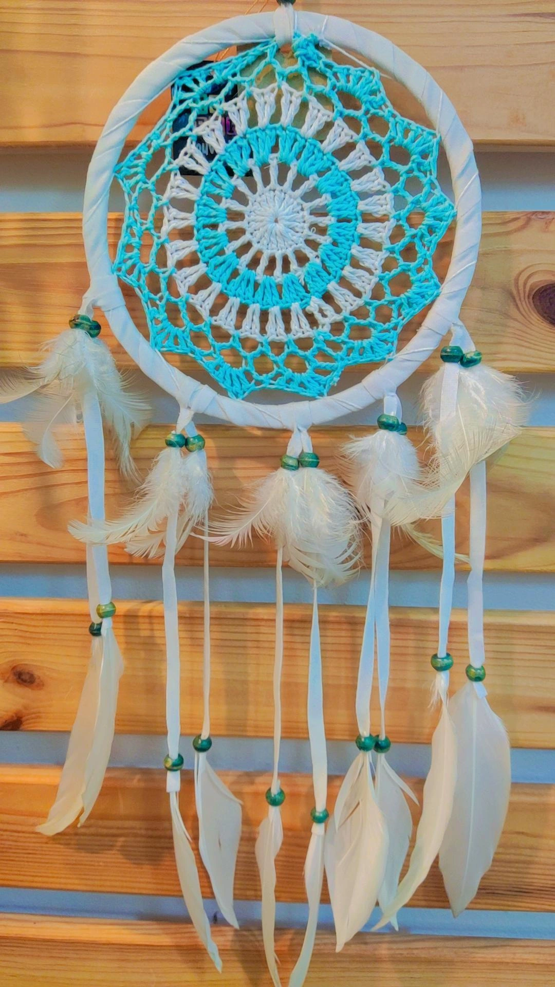 Apanhador de sonhos em crochet azul e branco com penas e contas verdes pendurado numa parede de madeira.