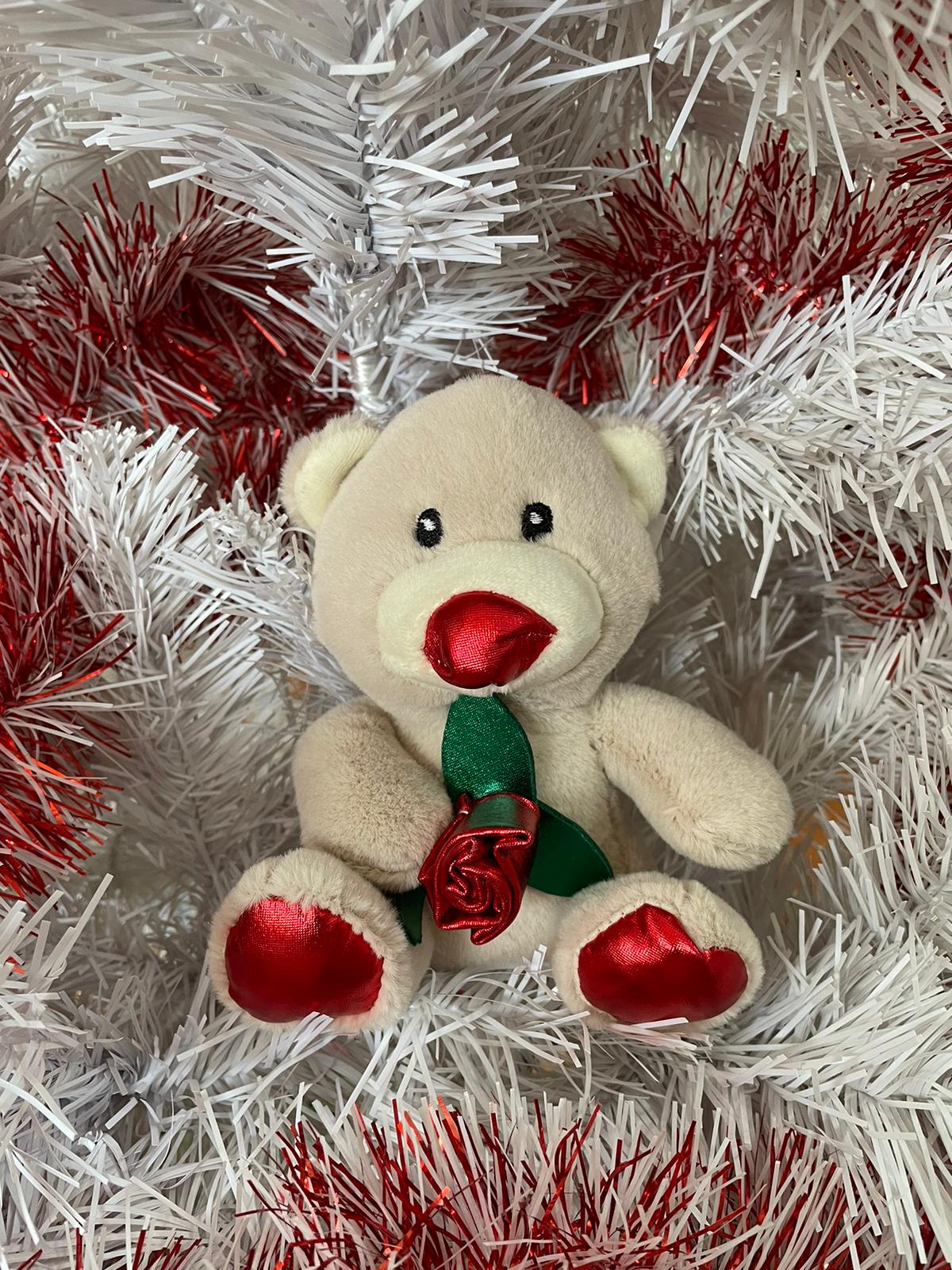 Ursinho de peluche bege com rosa vermelha segurada sentado em guirlanda branca e vermelha.