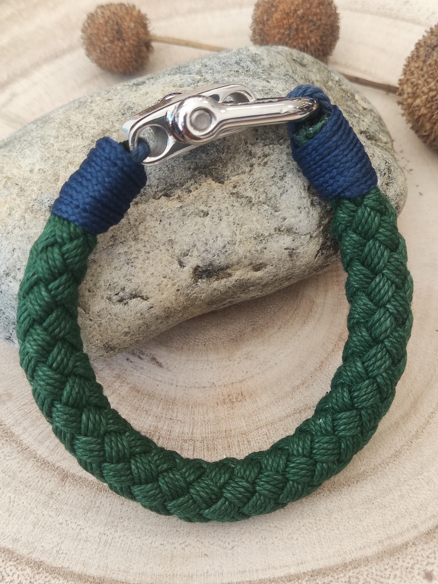 Pulseira trançada verde com fecho metálico prateado e detalhes azuis sobre pedra e madeira