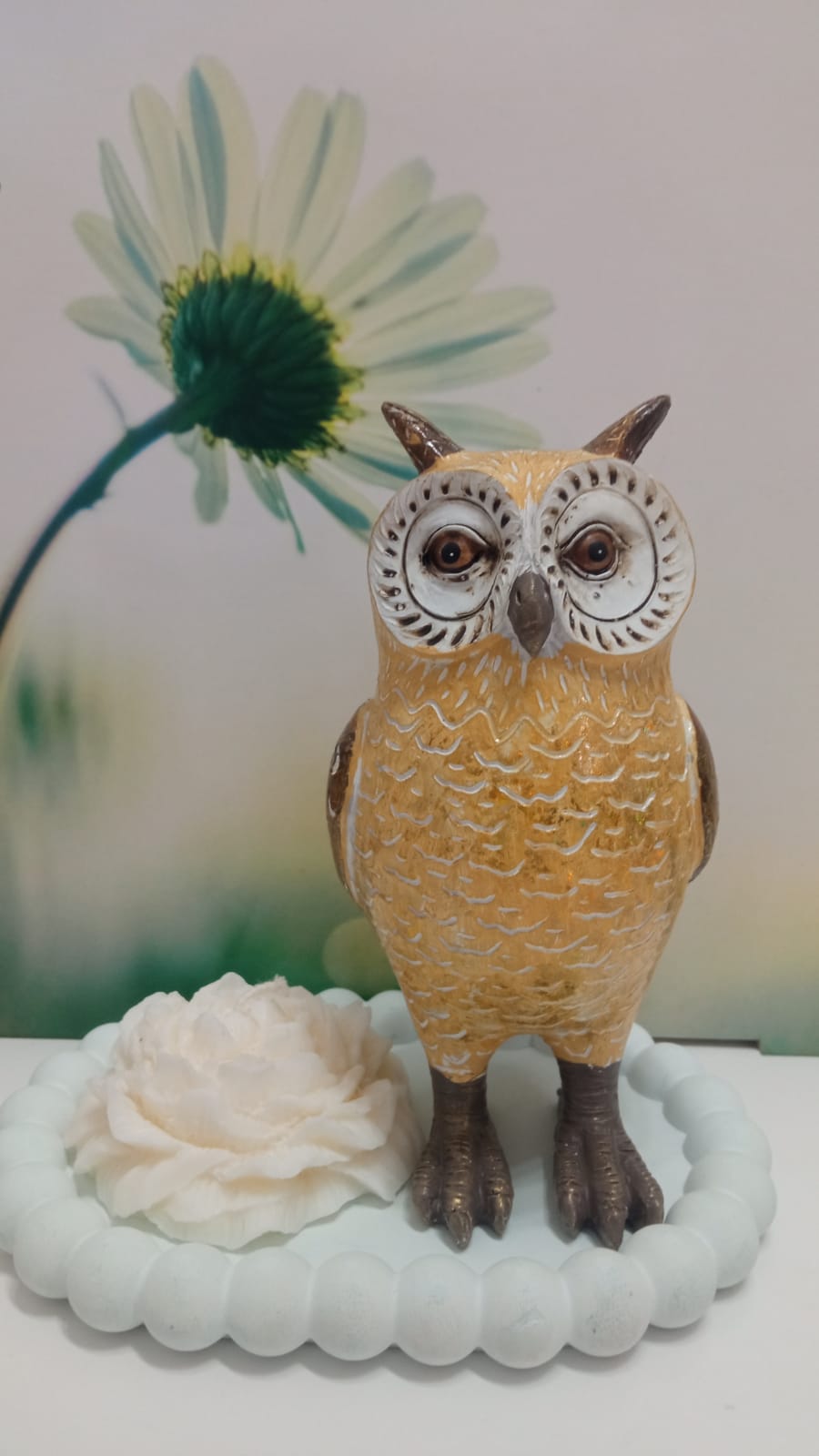 Estátua de coruja amarela e castanha em base branca com flor decorativa.