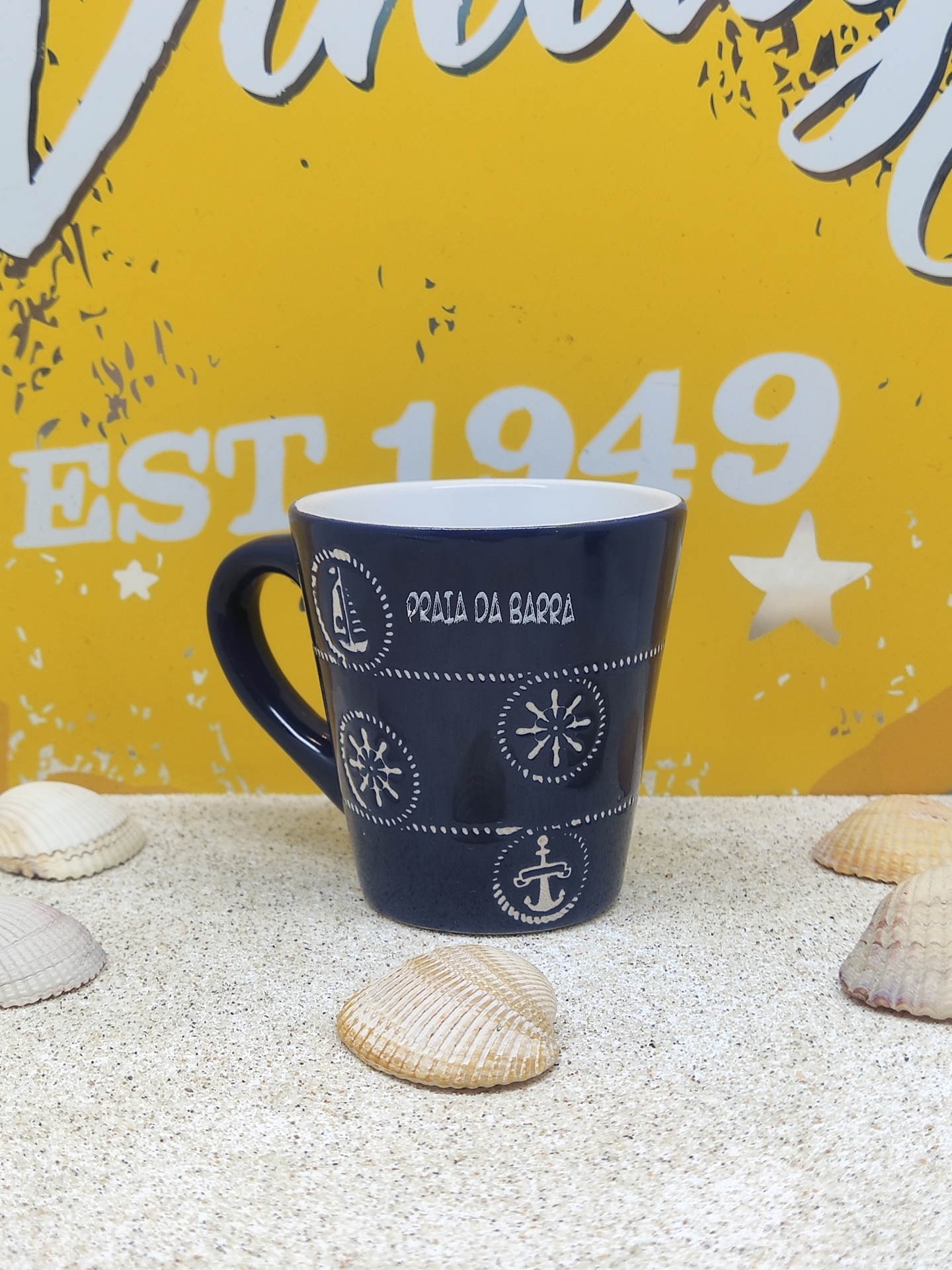 Caneca cerâmica azul com desenhos brancos marítimos e texto Praia da Barra sobre areia com conchas
