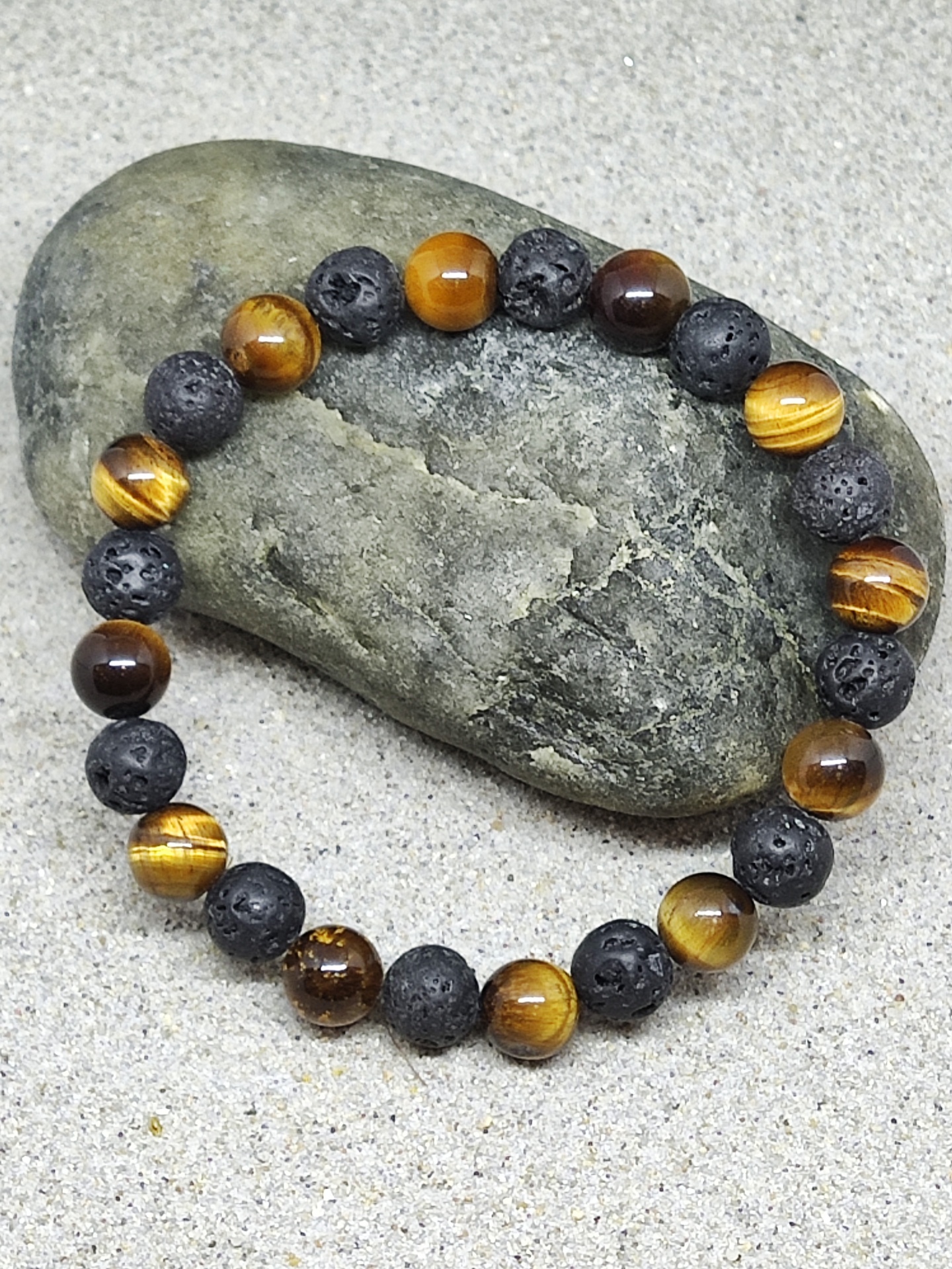 Pulseira com contas castanhas e negras porosas sobre pedra verde e fundo arenoso