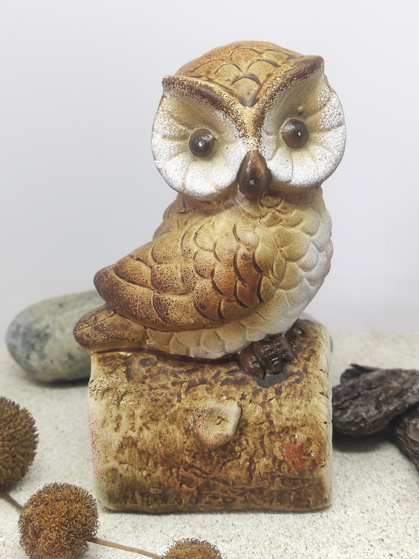 Figura decorativa de coruja em tons castanhos e beges sobre bloco que imita madeira.