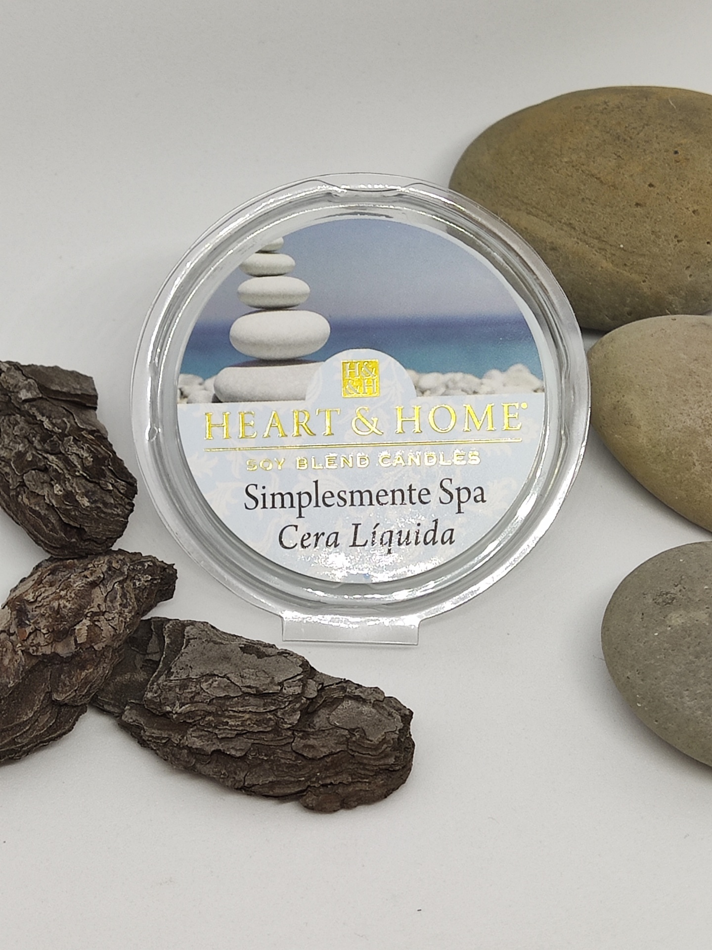 Vela em recipiente redondo transparente com tampa, texto 'Heart & Home Wax Blend Candles Simplesmente Spa Cera Líquida', rodeada por casca e pedras