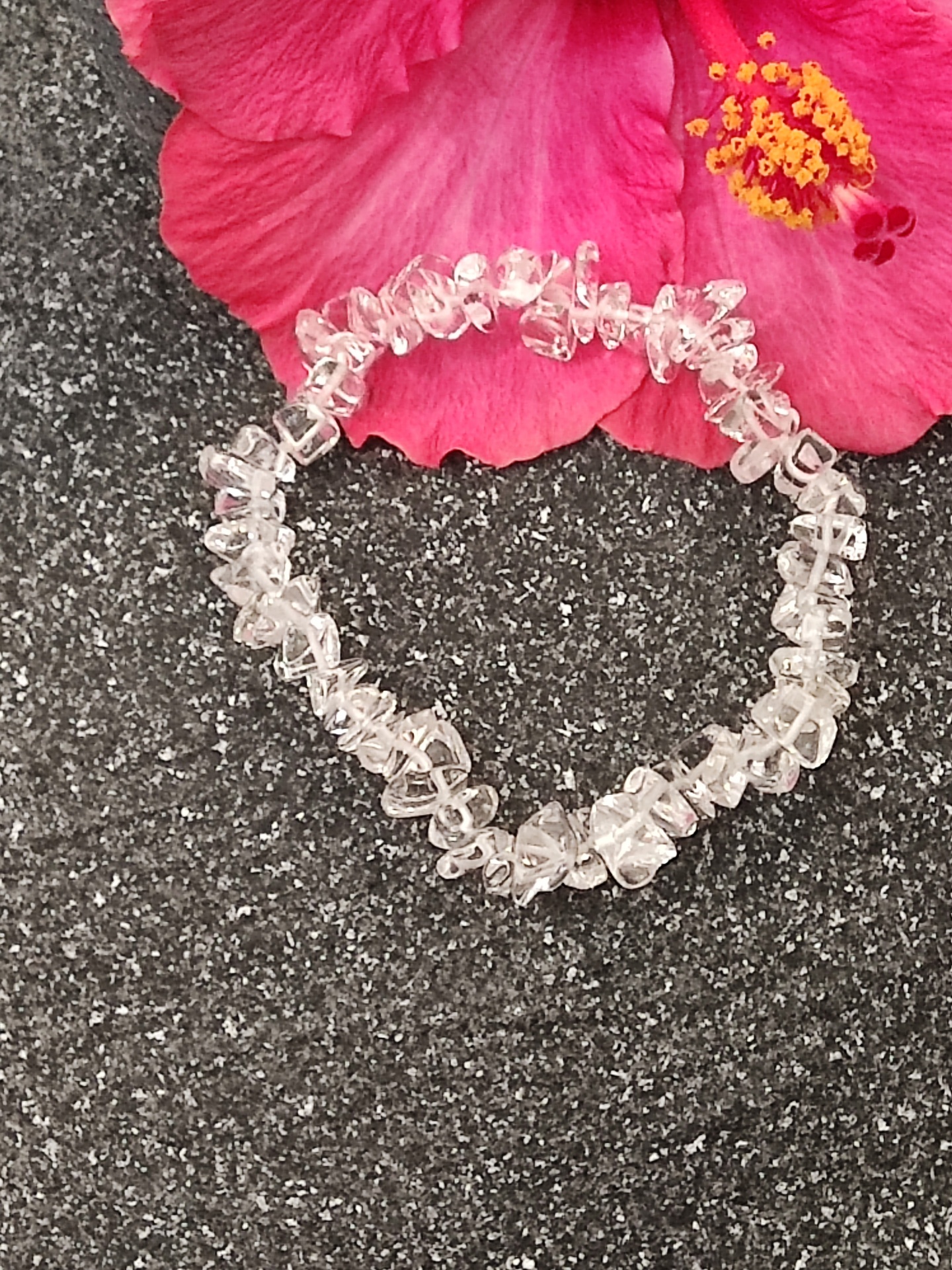 Pulseira de pedras transparentes sobre superfície preta ao lado de flor rosa